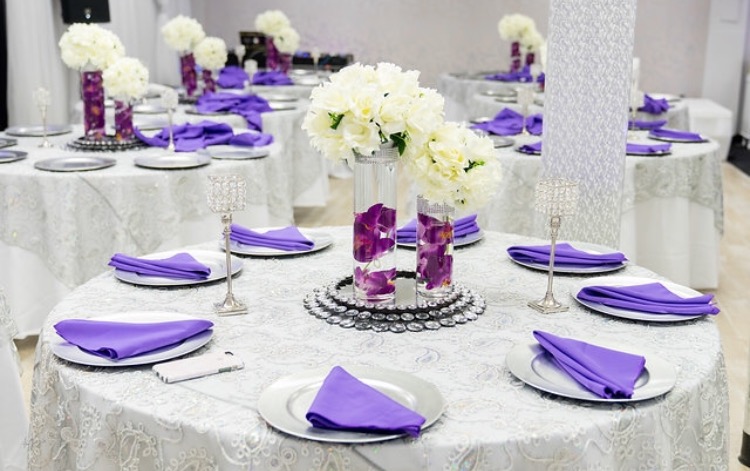 Elegant banquet hall set up for an event with round tables covered in white cloths, purple napkins, and floral centerpieces featuring white and purple flowers.