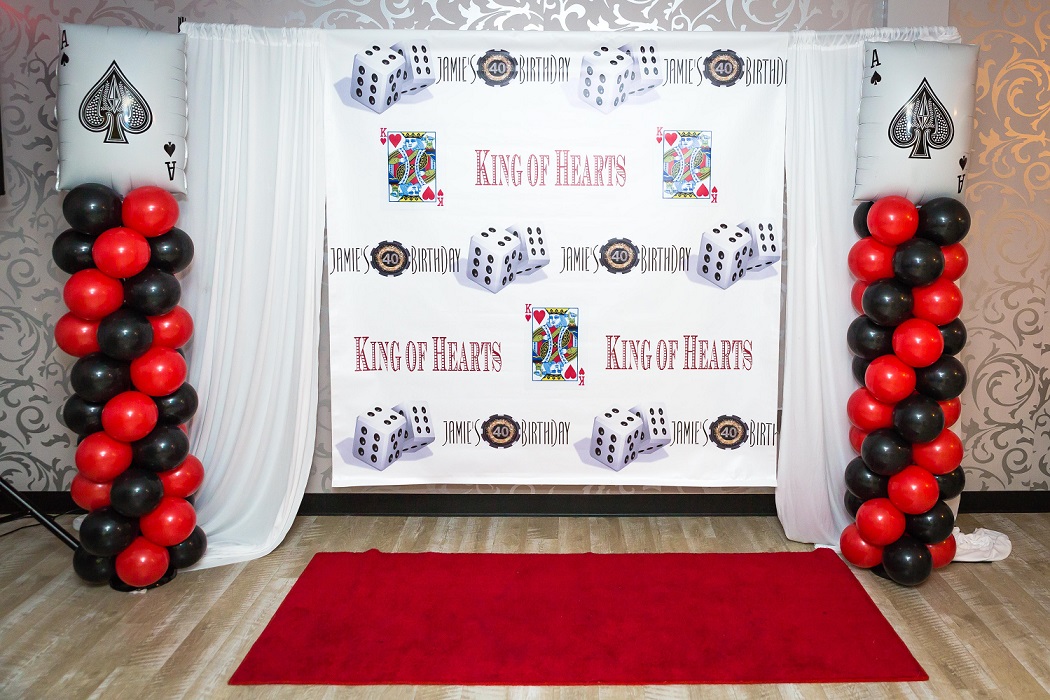 A casino-themed party backdrop with "king of hearts" and playing card illustrations, surrounded by black and red balloons and a red carpet in front.