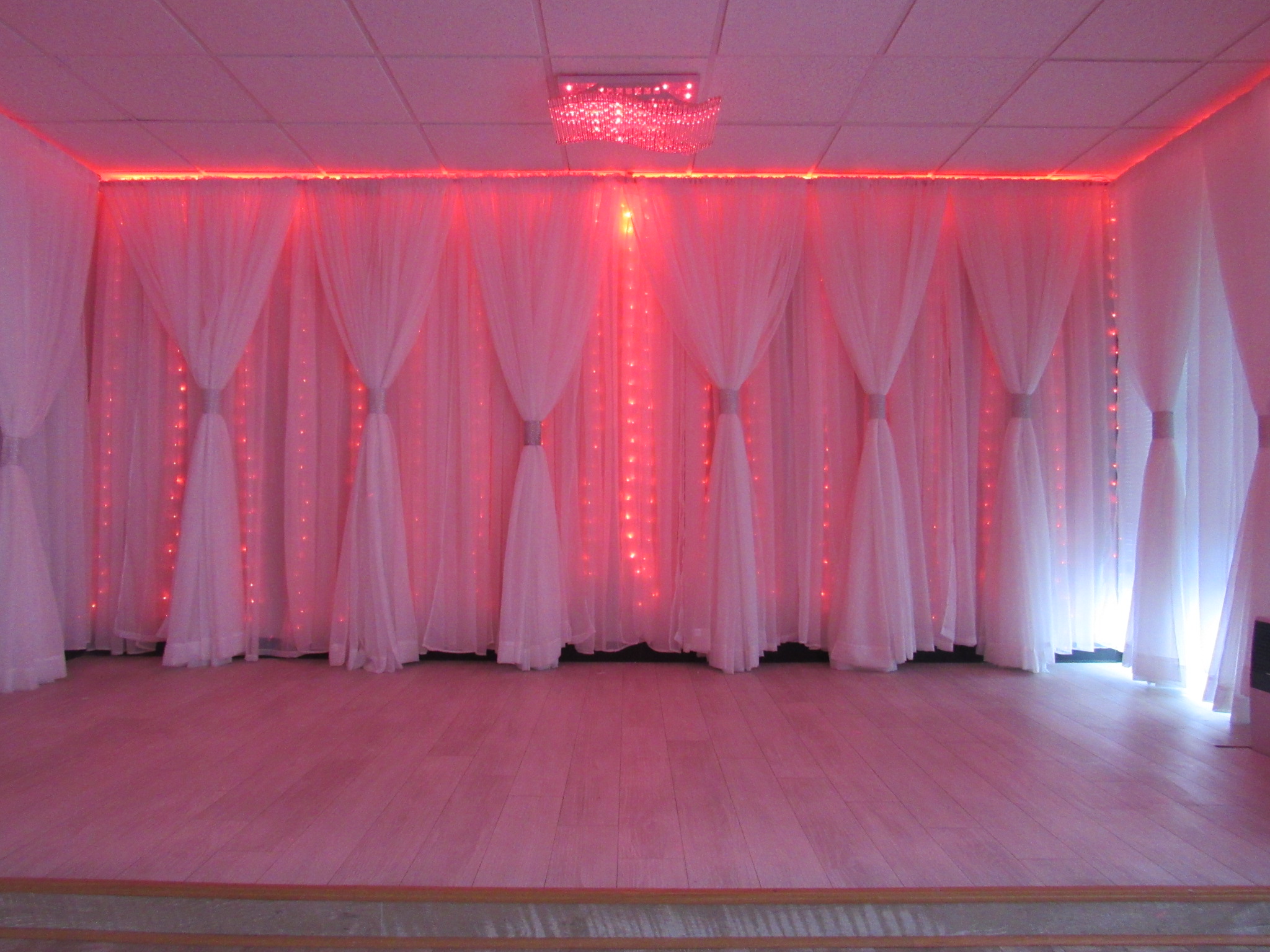 A warmly lit room with pink and red lights draped across sheer white curtains, highlighting an elegant wooden floor and a crystalline chandelier above.