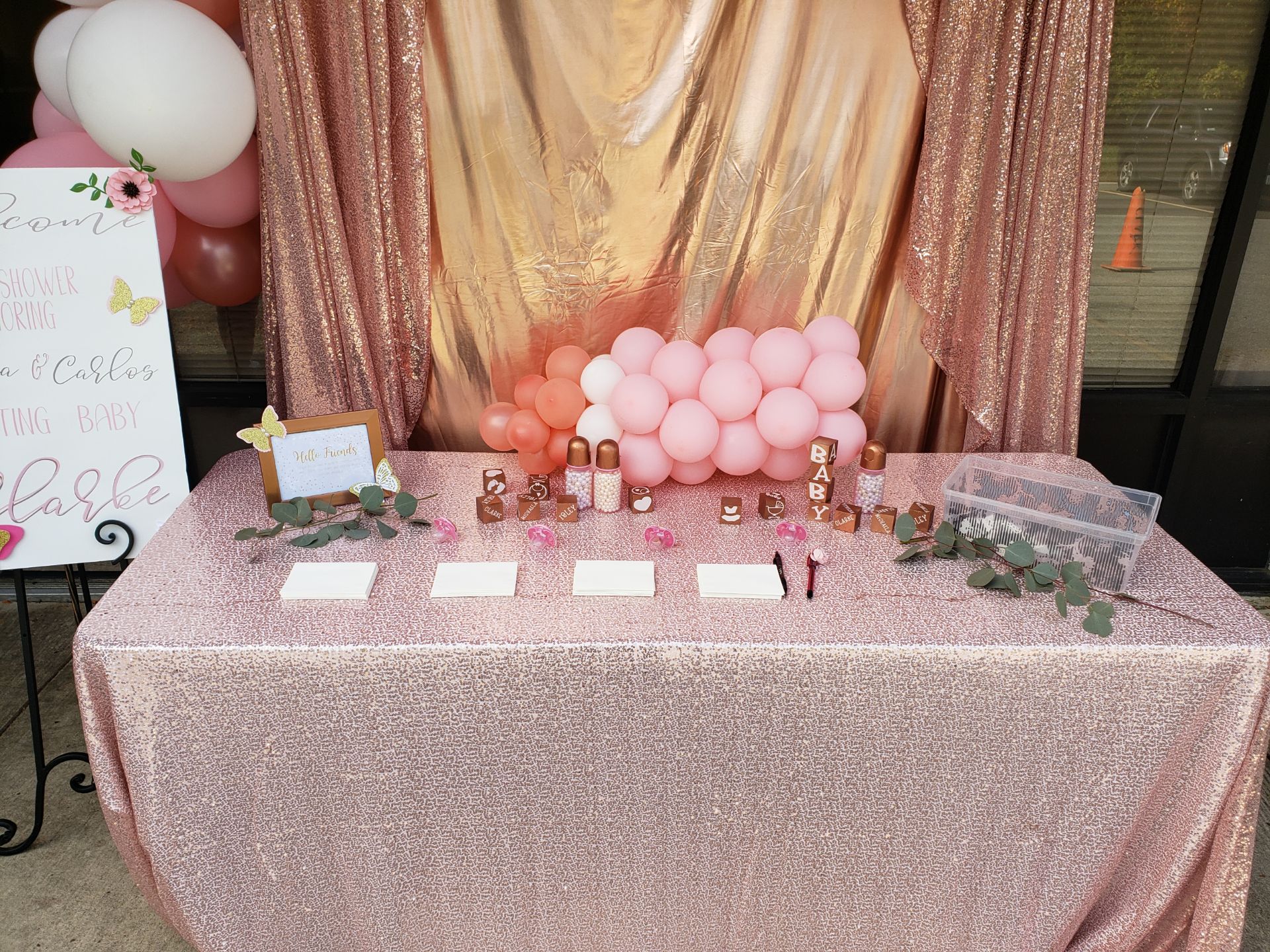 A baby shower setup with a pink and gold theme, featuring a table covered with a glittery cloth, decorated with balloons, a backdrop, and small boxes with labels.