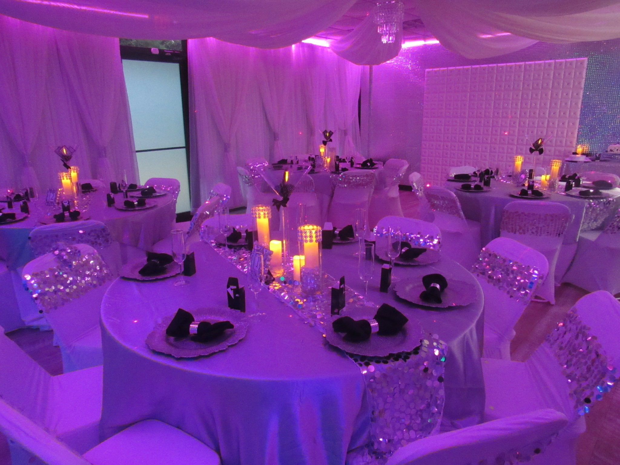 Elegant event setup with round tables covered in white linen, adorned with black napkins and glowing centerpieces. the room features pink lighting and draped white curtains.