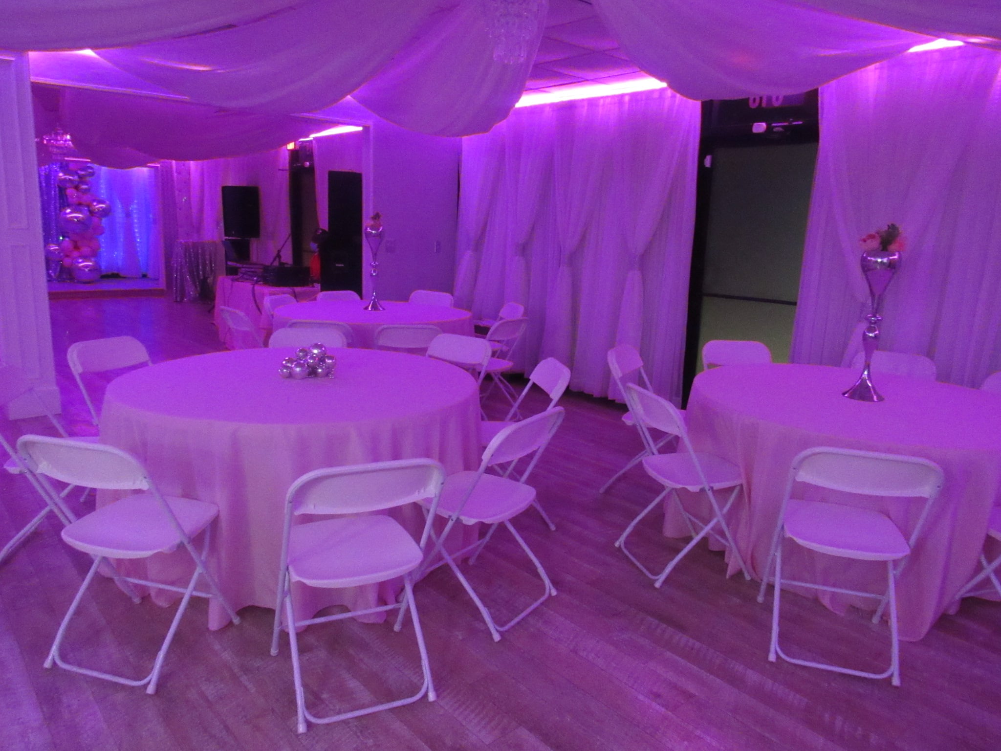 An event hall lit by purple lighting with round tables covered in white cloths, white chairs, and floral centerpieces. white drapes and a dance floor are visible in the background.