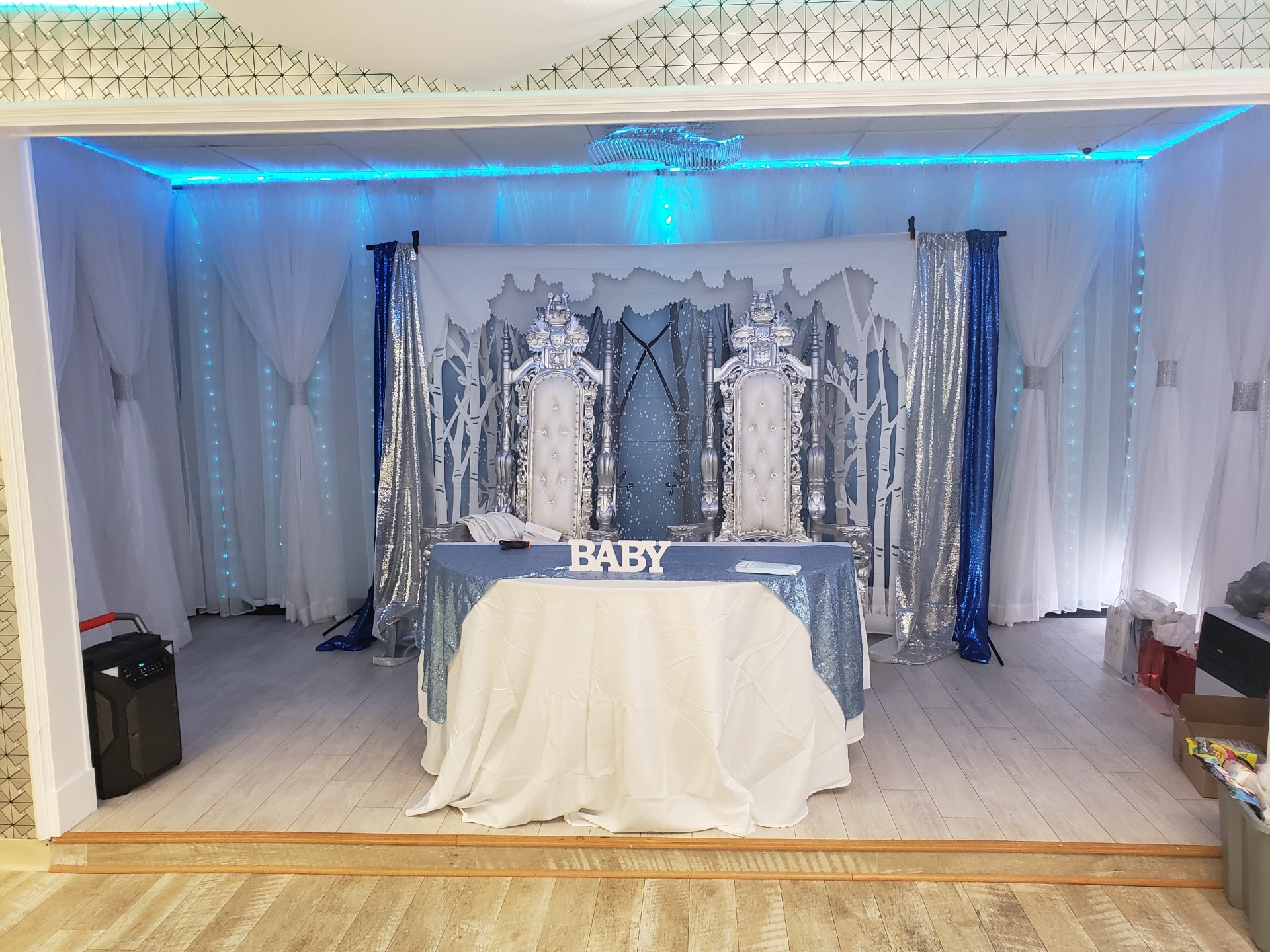 Elegant baby shower setup with two ornate silver chairs under a blue-tinted light, white and blue drapery backdrop, crystal accents, and a table with a 'baby' sign.