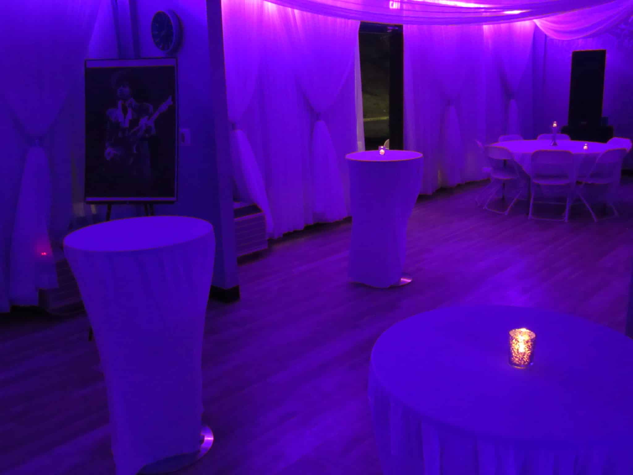 An event space illuminated by blue lights featuring round tables with white tablecloths, a poster of a guitar player, and draped white curtains.