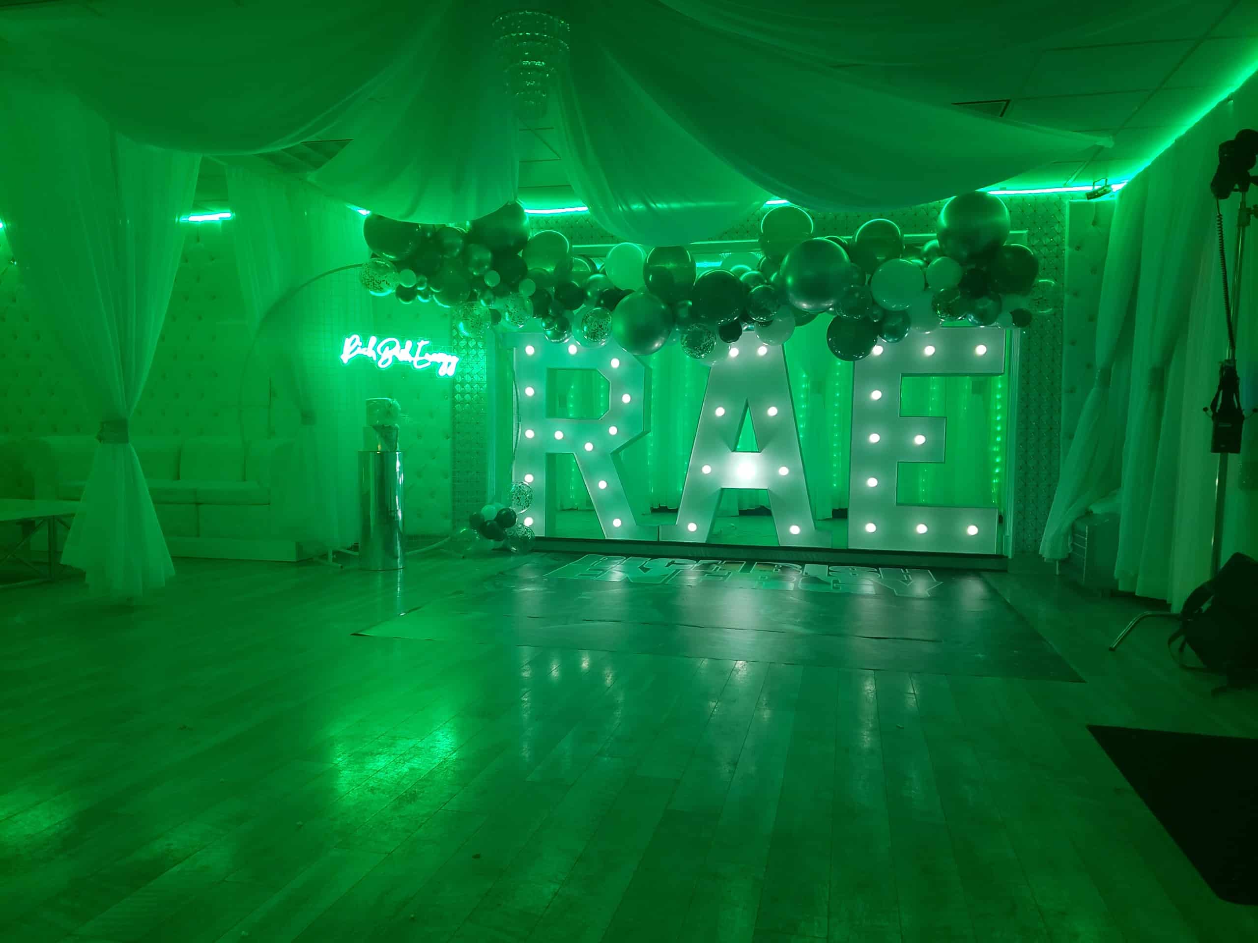 An event space illuminated in green light with a large, lit-up "rae" sign decorated with balloons and a neon sign that reads "bright." white drapery and a chandelier adorn the background.