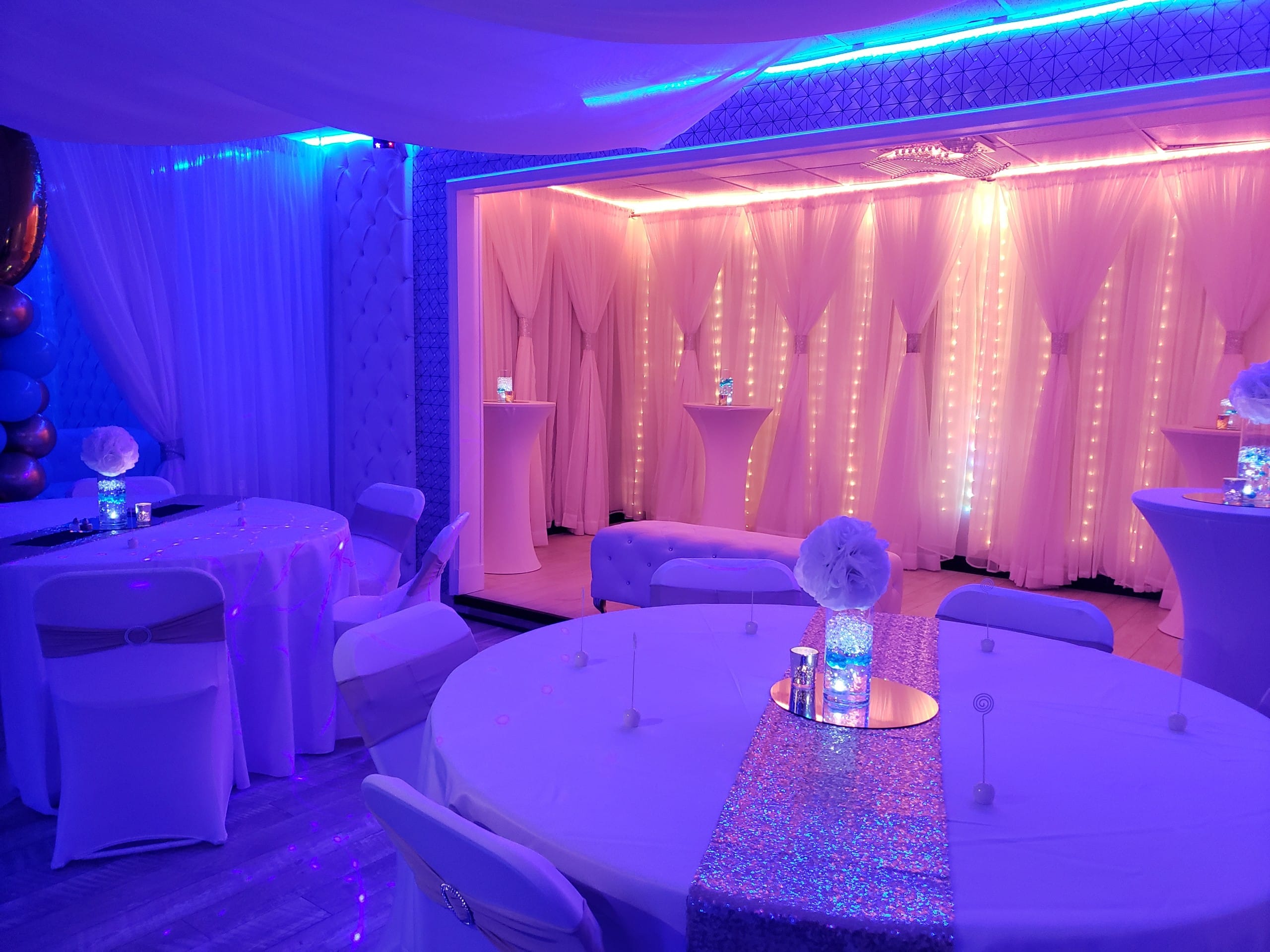 Elegant event space decorated with blue and pink lighting, featuring round tables with white tablecloths and centerpieces, surrounded by draped curtains and illuminated accents.
