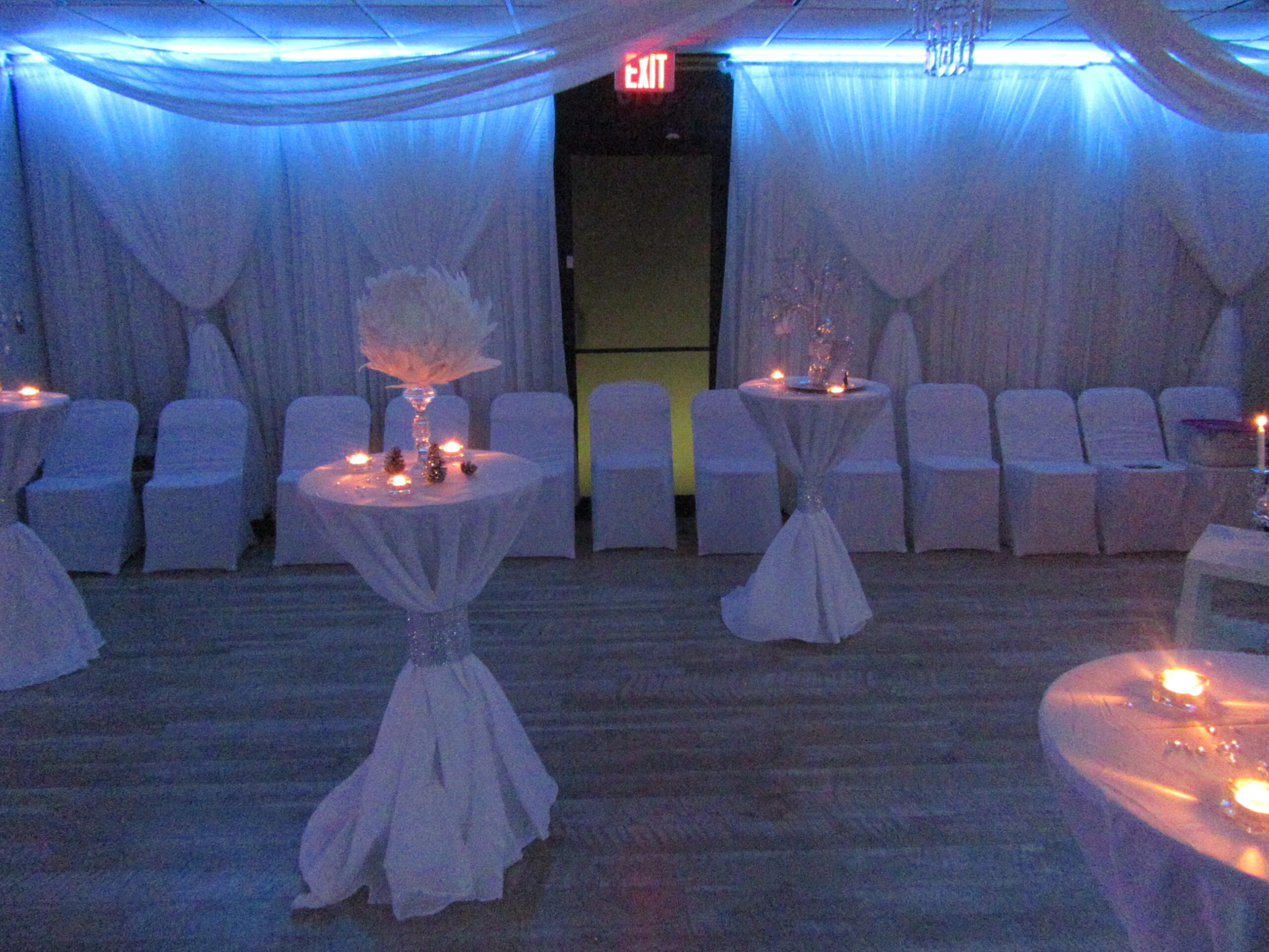 A dimly lit event room with draped white linens, round tables covered with white cloths, and chairs wrapped in white with centerpieces featuring lit candles and floral decorations.
