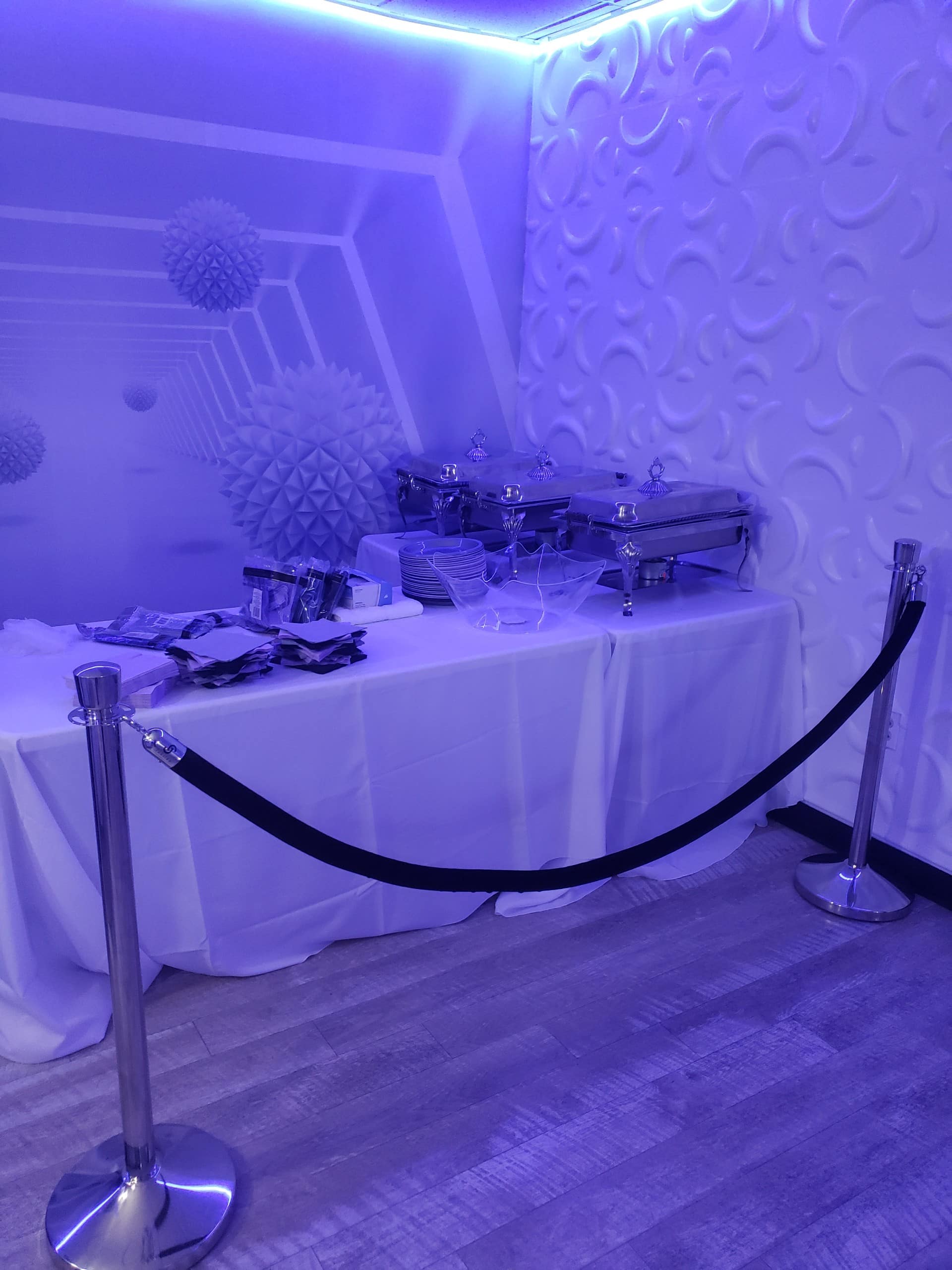 A buffet table set up in a room with purple lighting, featuring chafing dishes and serving utensils, secured by a black velvet rope barrier.
