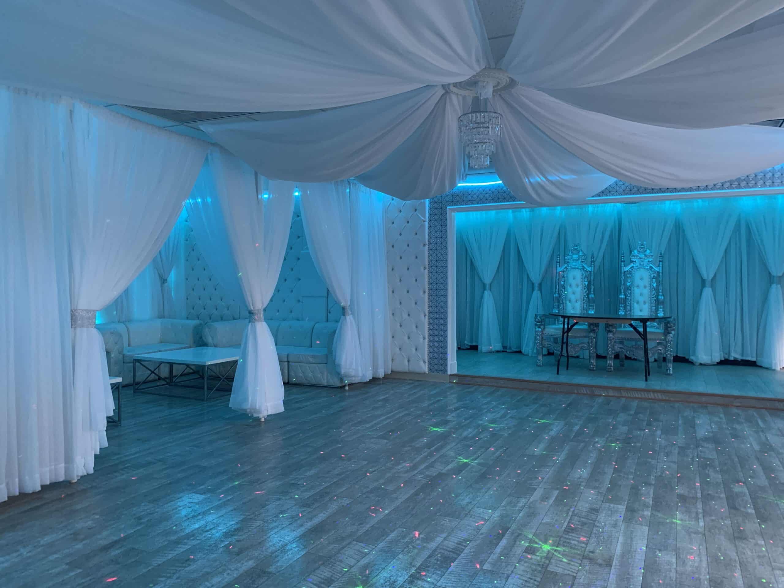 Elegant event space decorated in white and blue, featuring draped ceilings, white sheer curtains, and sitting areas with luxurious couches. soft blue lighting adds a serene atmosphere.