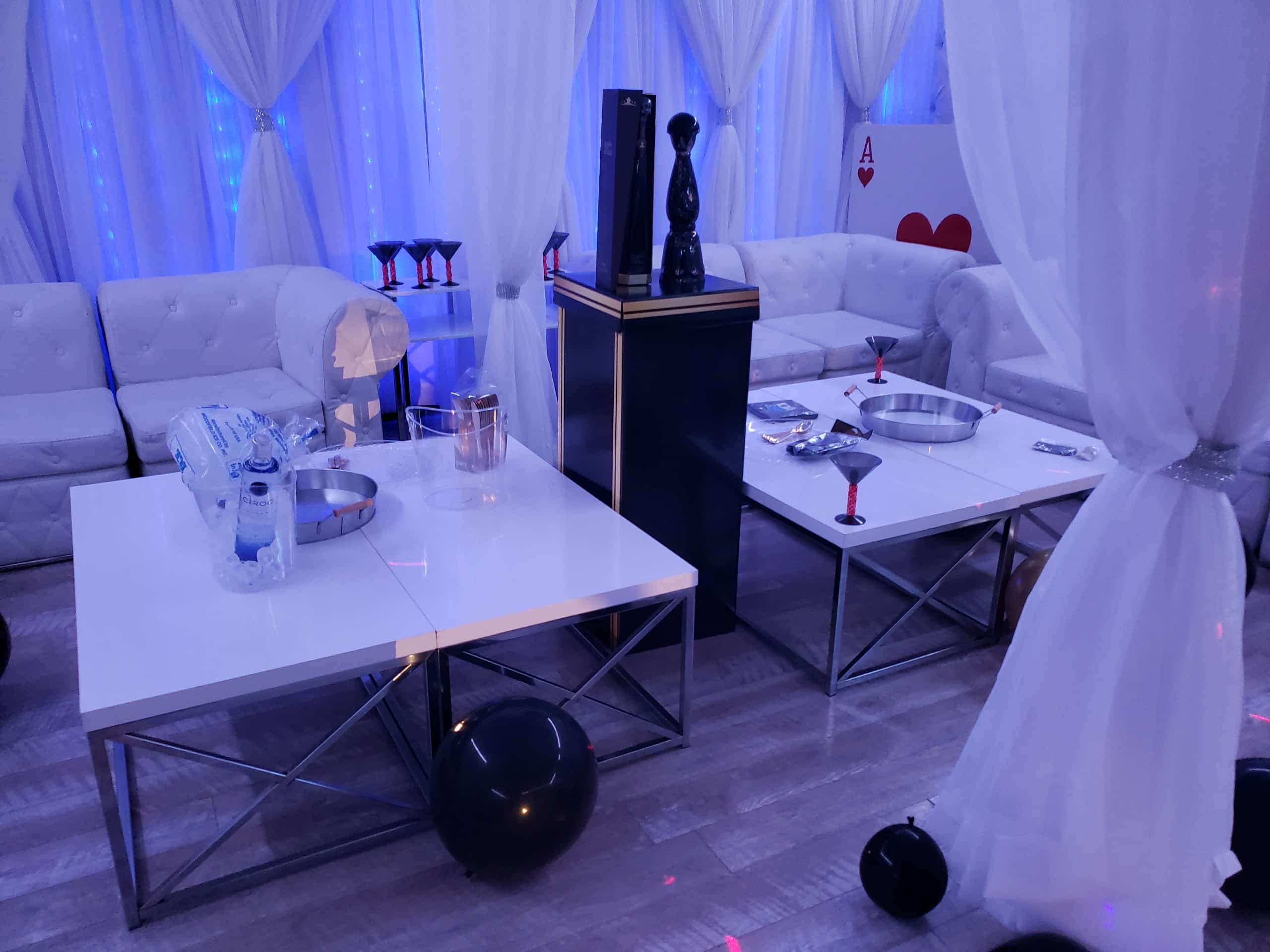 An elegant party setting with white sofas, translucent curtains, and tables decorated with black balloons, glassware, and white serving dishes. soft blue lighting adds to the ambiance.
