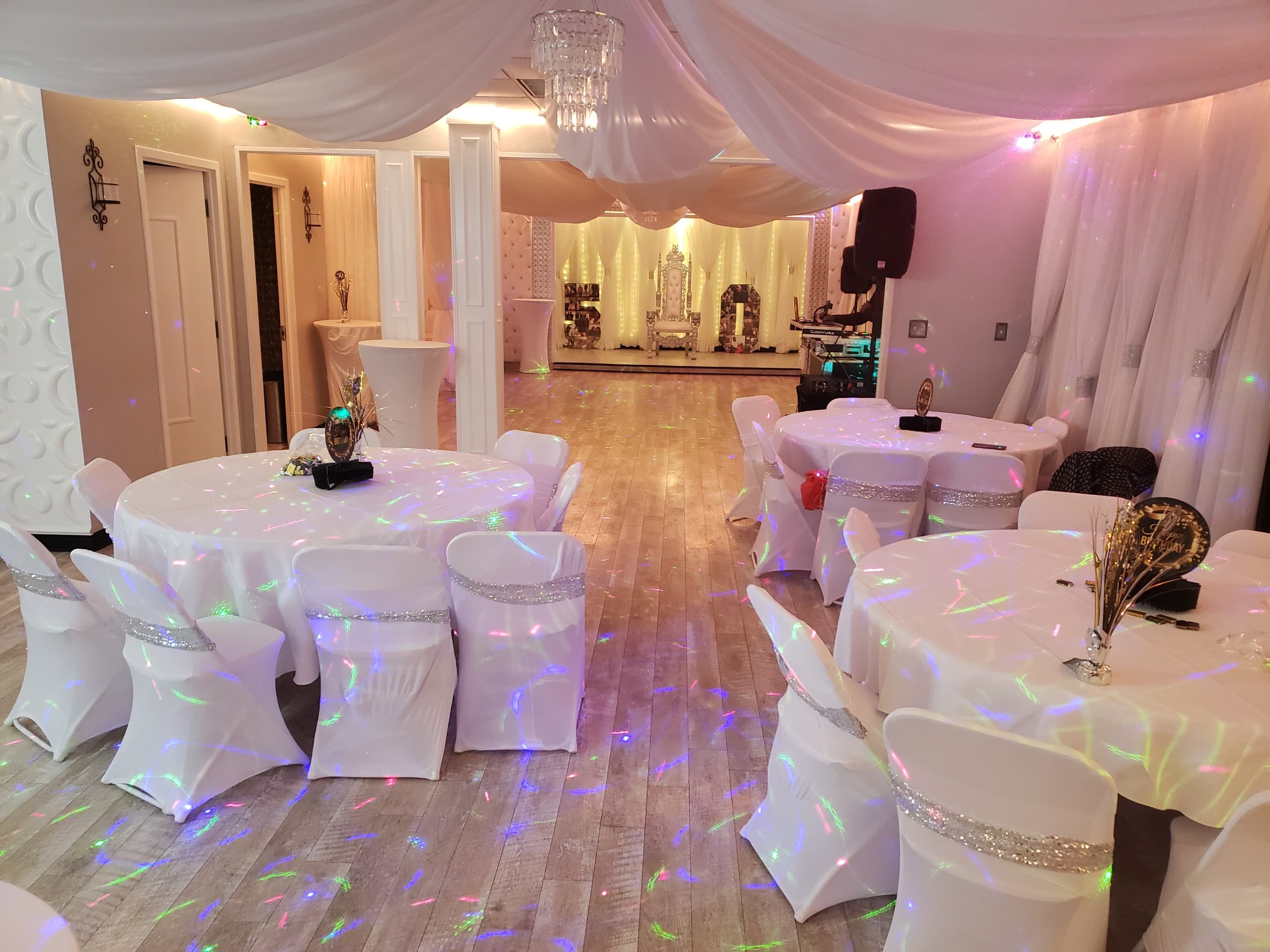 An elegantly decorated event hall with white draped tables, sparkling table settings, colorful floor lights, and a large chandelier, creating a festive and sophisticated ambiance.