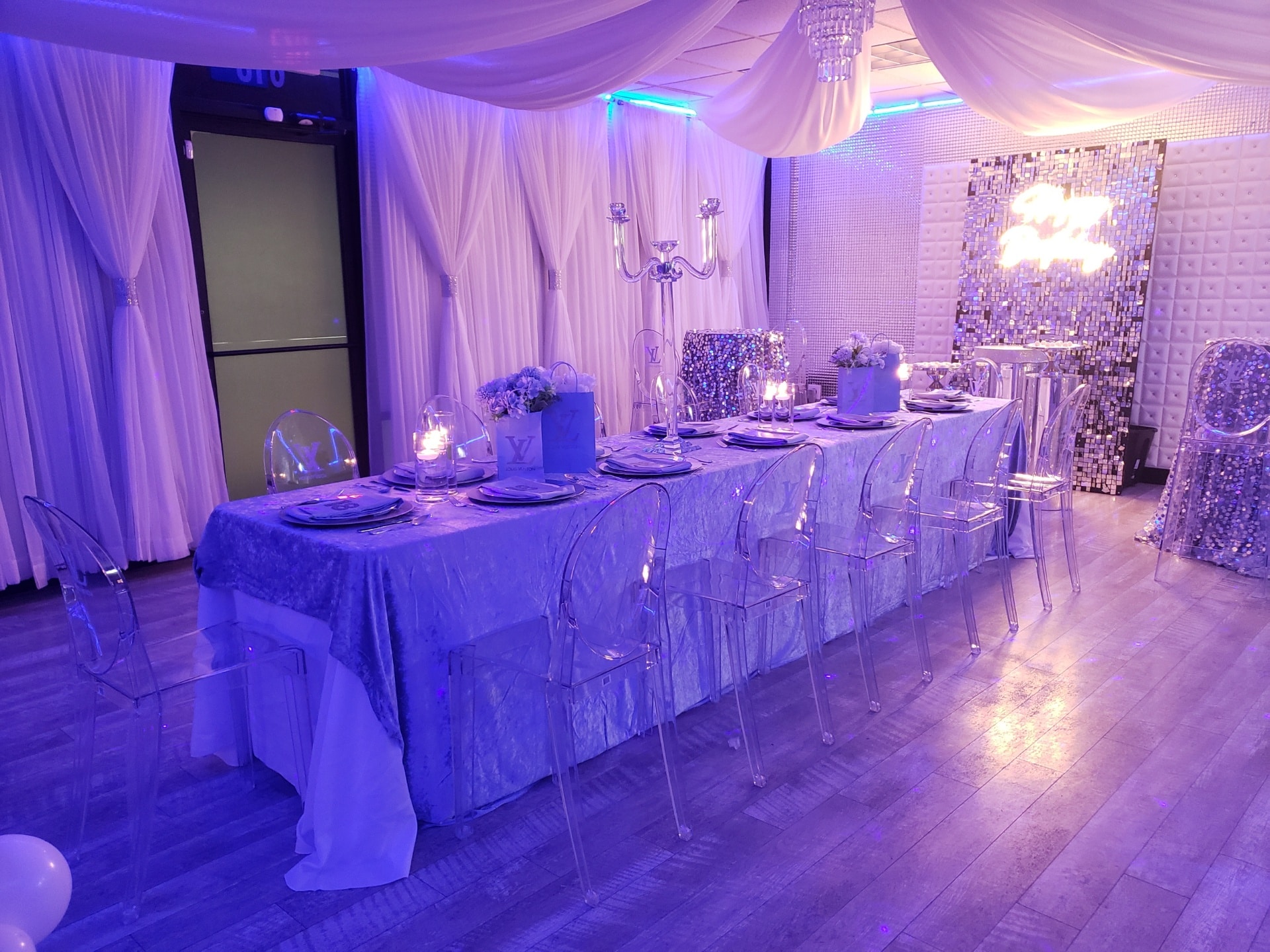 Elegant event setting with a long table covered in a blue shimmering cloth, transparent chairs, and silver candelabras, adorned by soft purple lighting and white drapery.