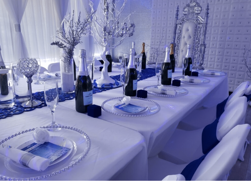 Elegant dining setup in a room with blue lighting, featuring a long table set with white plates, silver cutlery, crystal glasses, and champagne bottles, highlighted by white and silver decorative elements.