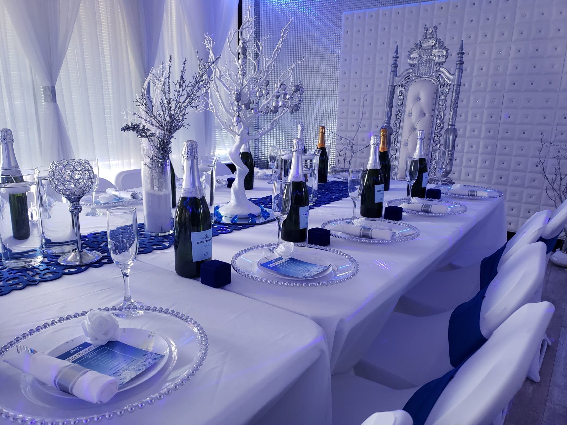 An elegant event setup featuring a long white table with ornate silver and white decorations, champagne bottles, and fine dining place settings under blue lighting.
