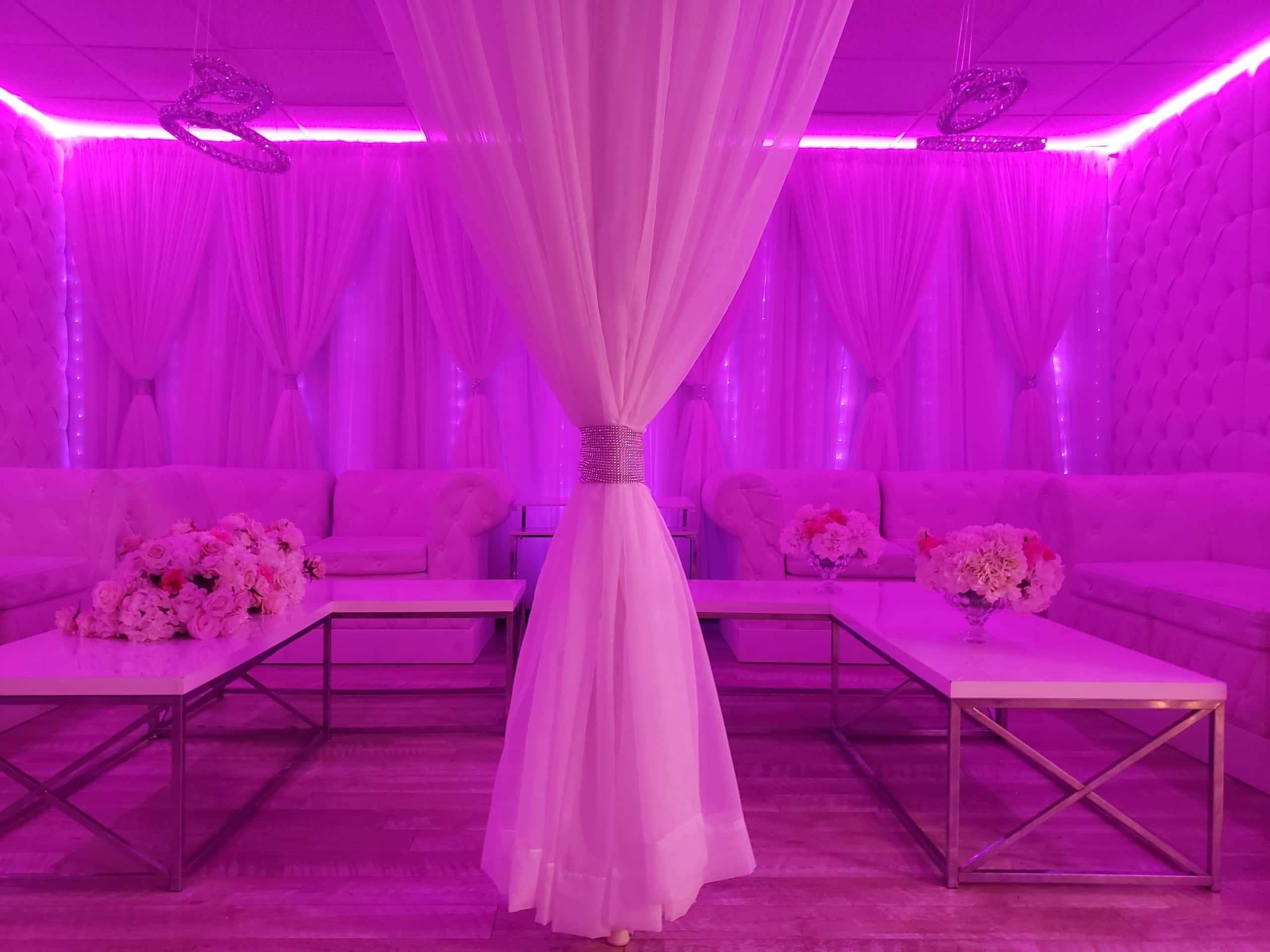 A lavish event space with pink lighting, featuring elegant white sofas, crystal chandeliers, floral arrangements, and draped white curtains.