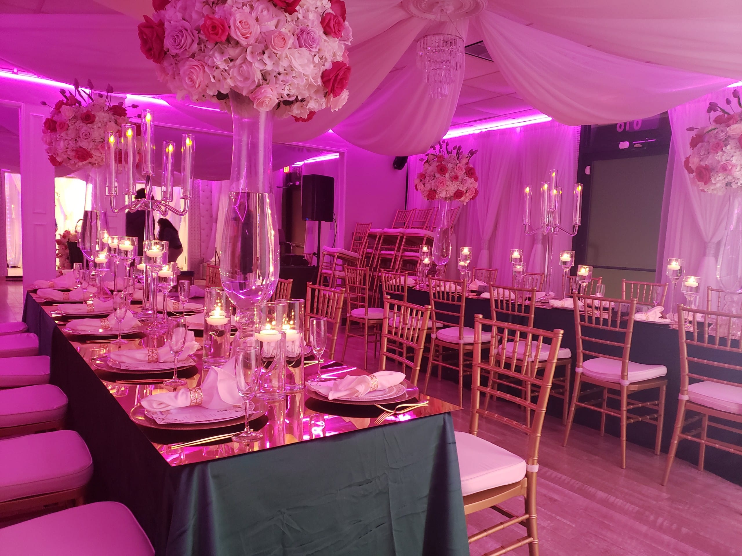 Elegant wedding reception setup featuring long tables with white linens, gold chairs, tall floral centerpieces, and pink ambient lighting.