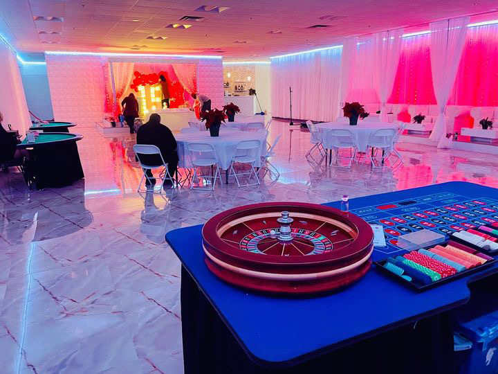A spacious room set up for an event, featuring a roulette table in the foreground, round tables with chairs, and a stage with red lighting in the background.