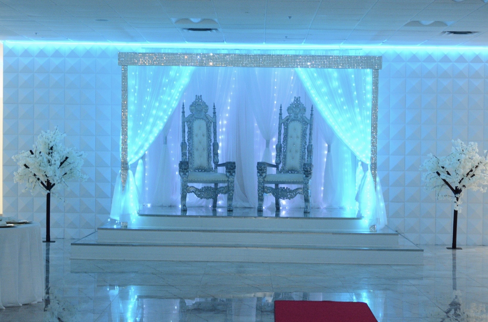 Elaborate wedding stage with two ornate chairs set against a backlit blue curtain, flanked by white decorative trees, in a spacious, well-lit hall.