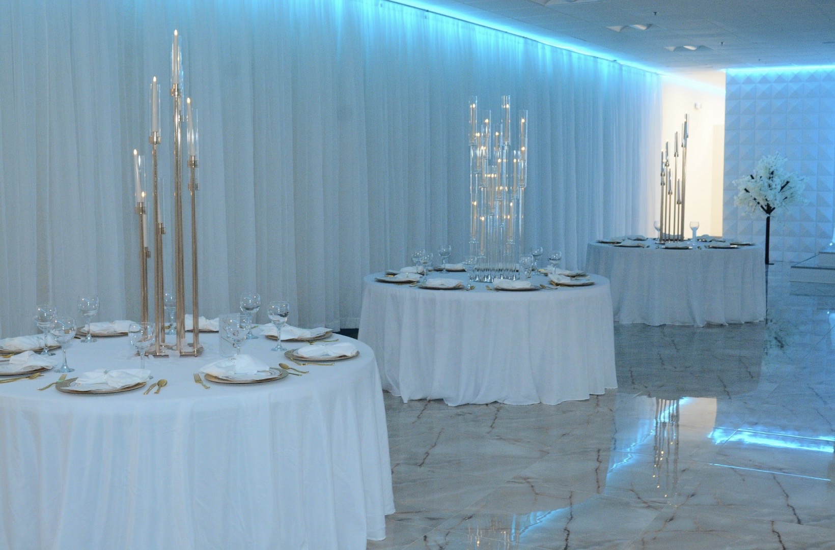 Elegant banquet hall setup with tables adorned with white tablecloths, gold rimmed plates, and crystal glassware. tall candle stands and a serene blue lighting backdrop enhance the luxurious ambiance.