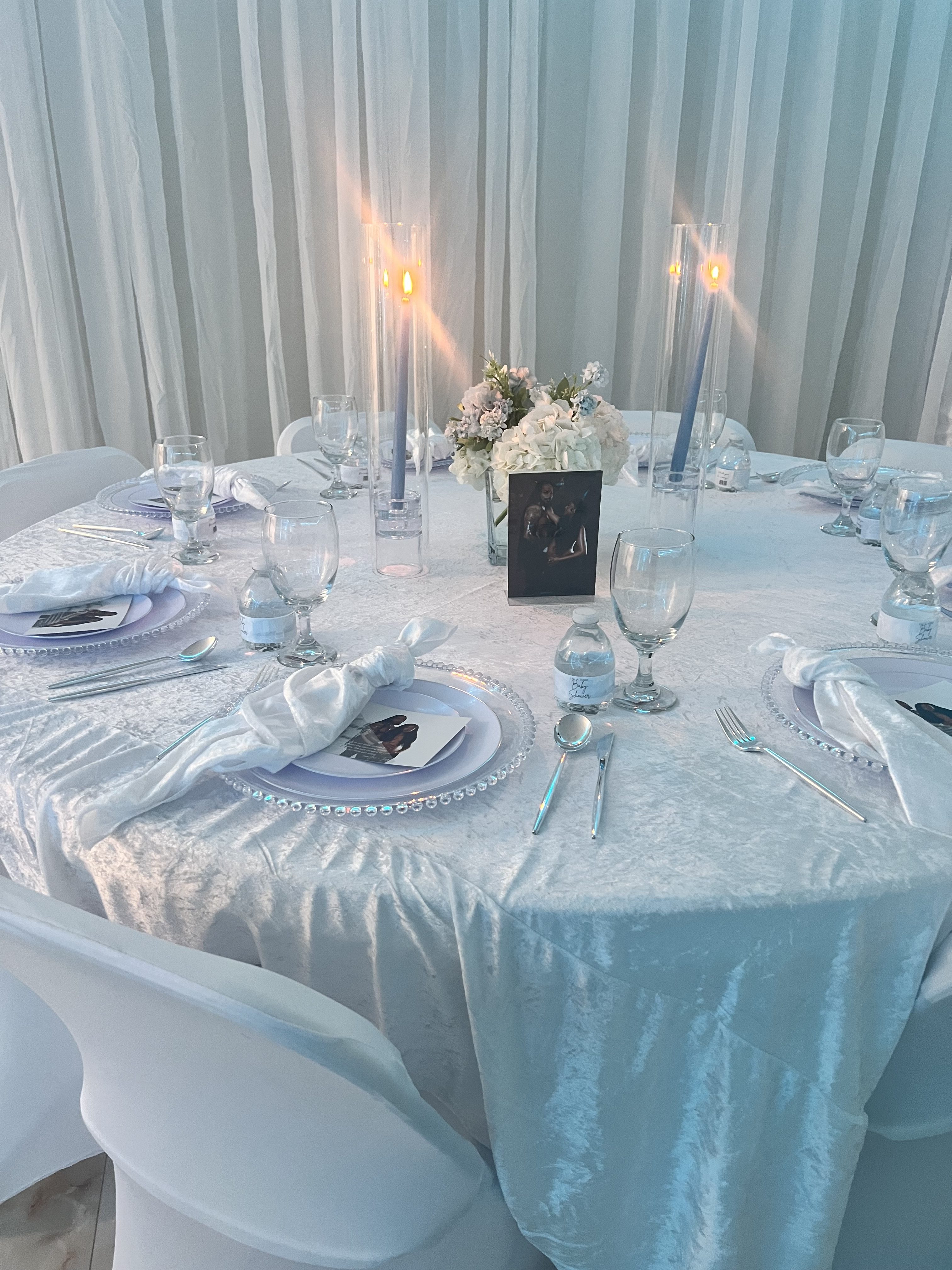 Elegant dining setting for a wedding reception with a white theme, featuring a round table with floral centerpieces, crystal candlesticks, and place settings with a photo frame.