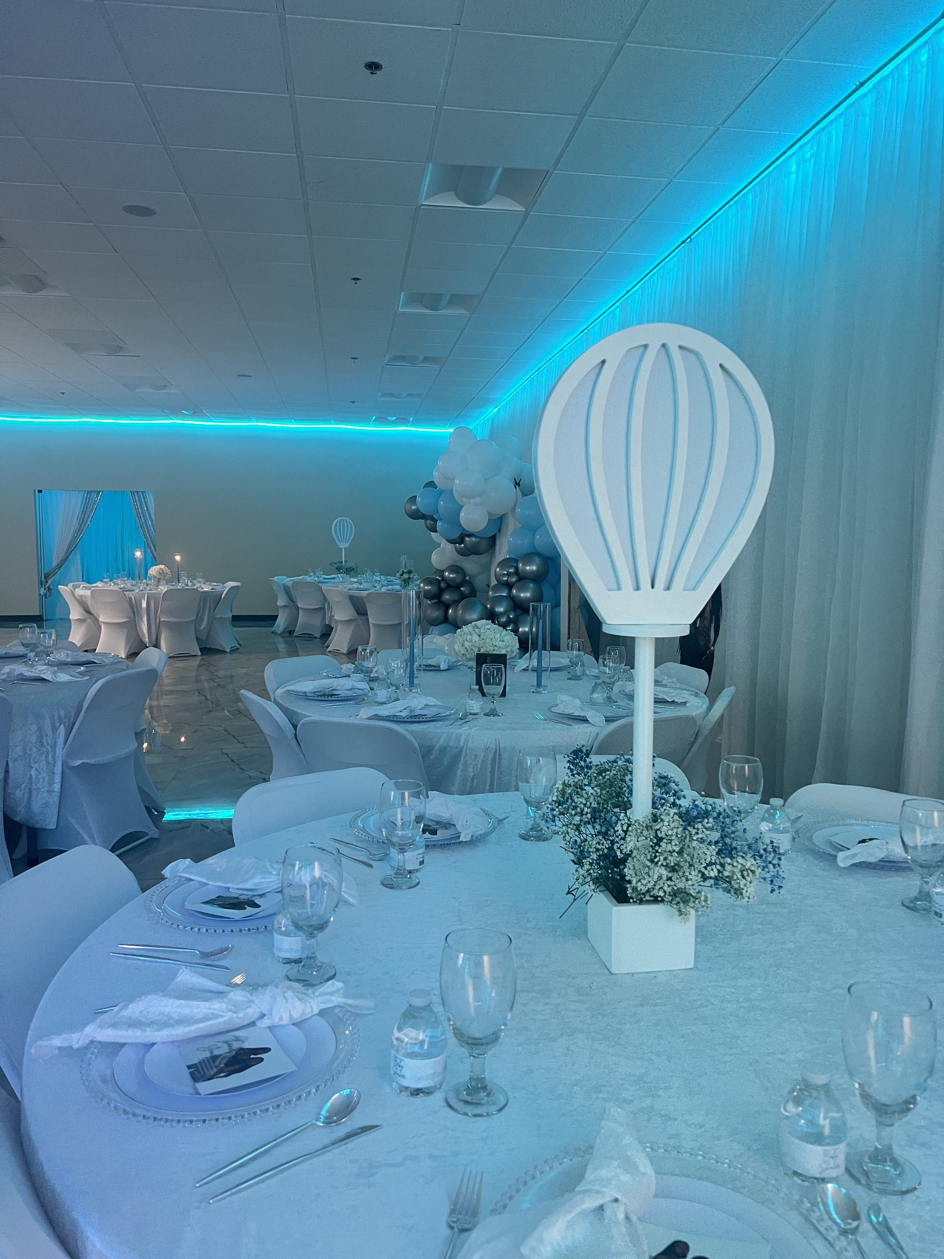 An elegantly decorated event venue with a blue theme, featuring round tables set with white linens, glasses, and plates. balloon sculptures and blue lighting add to the festive atmosphere.