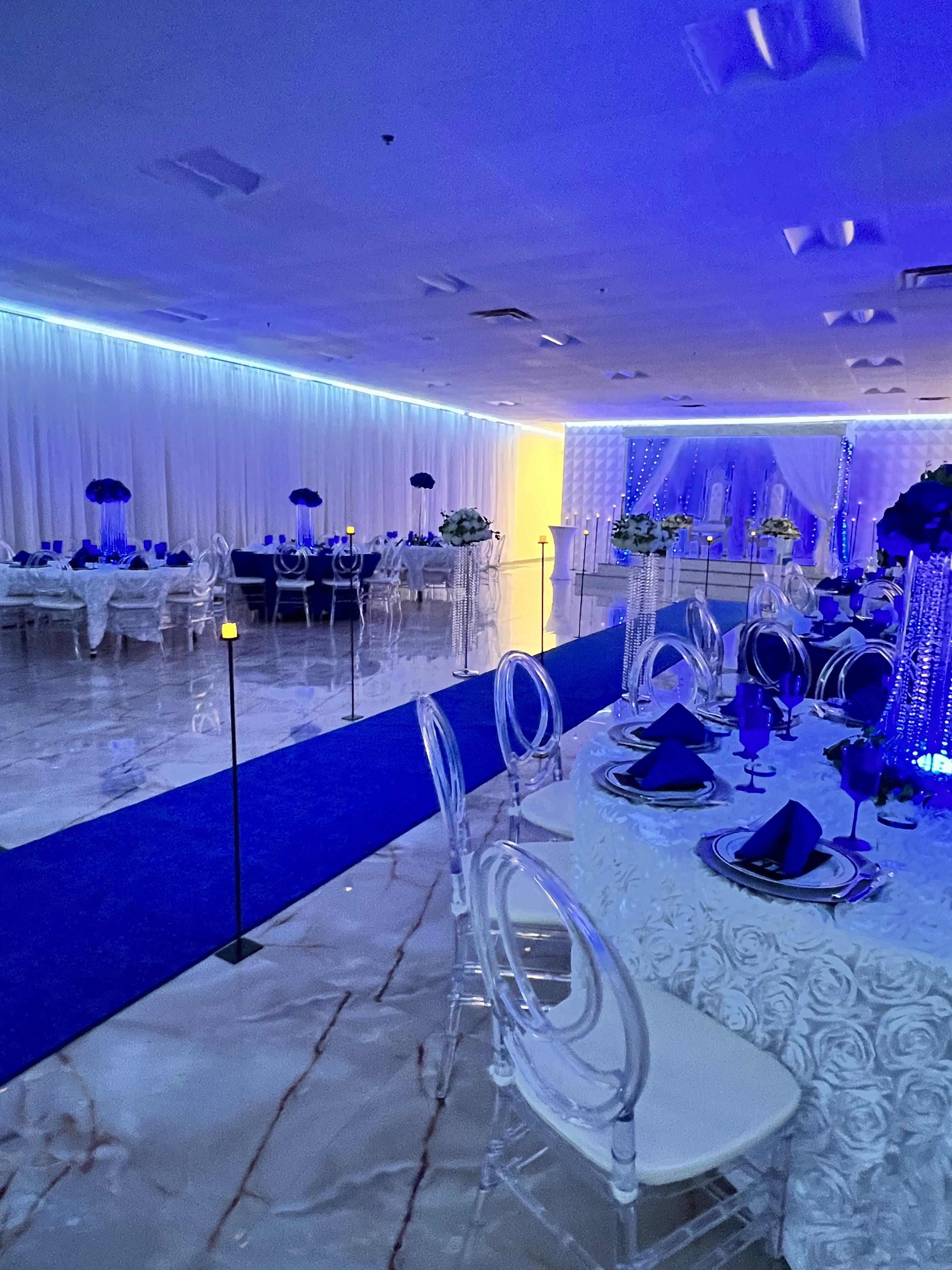 Elegant banquet hall decorated for an event, featuring a blue light ambiance, white and blue table settings on marble floors, and draped ceilings with blue uplighting.