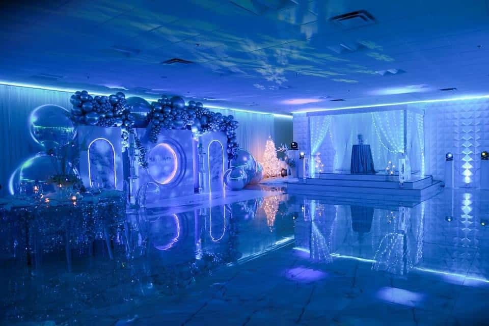 Elegant event space illuminated in blue light, featuring balloon decorations, reflective floor, and a central podium with curtains. a modern, sophisticated setting for a celebration.