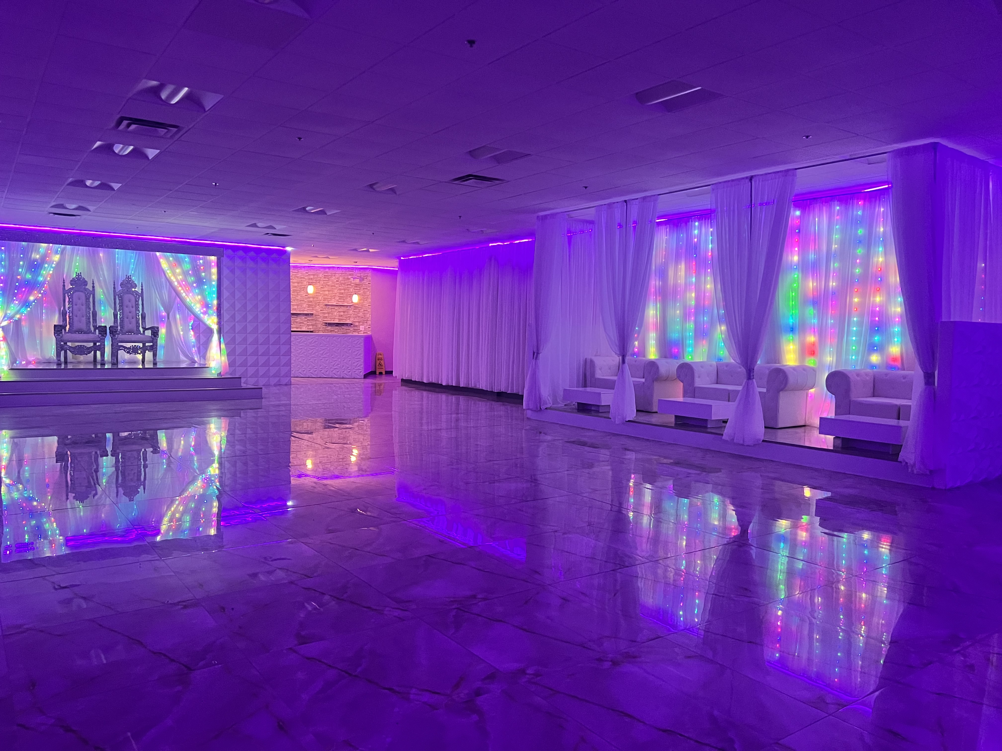 An event space with glossy marble floors reflecting colorful led lights from wall panels, white drapery, and a decorated stage with two chairs.
