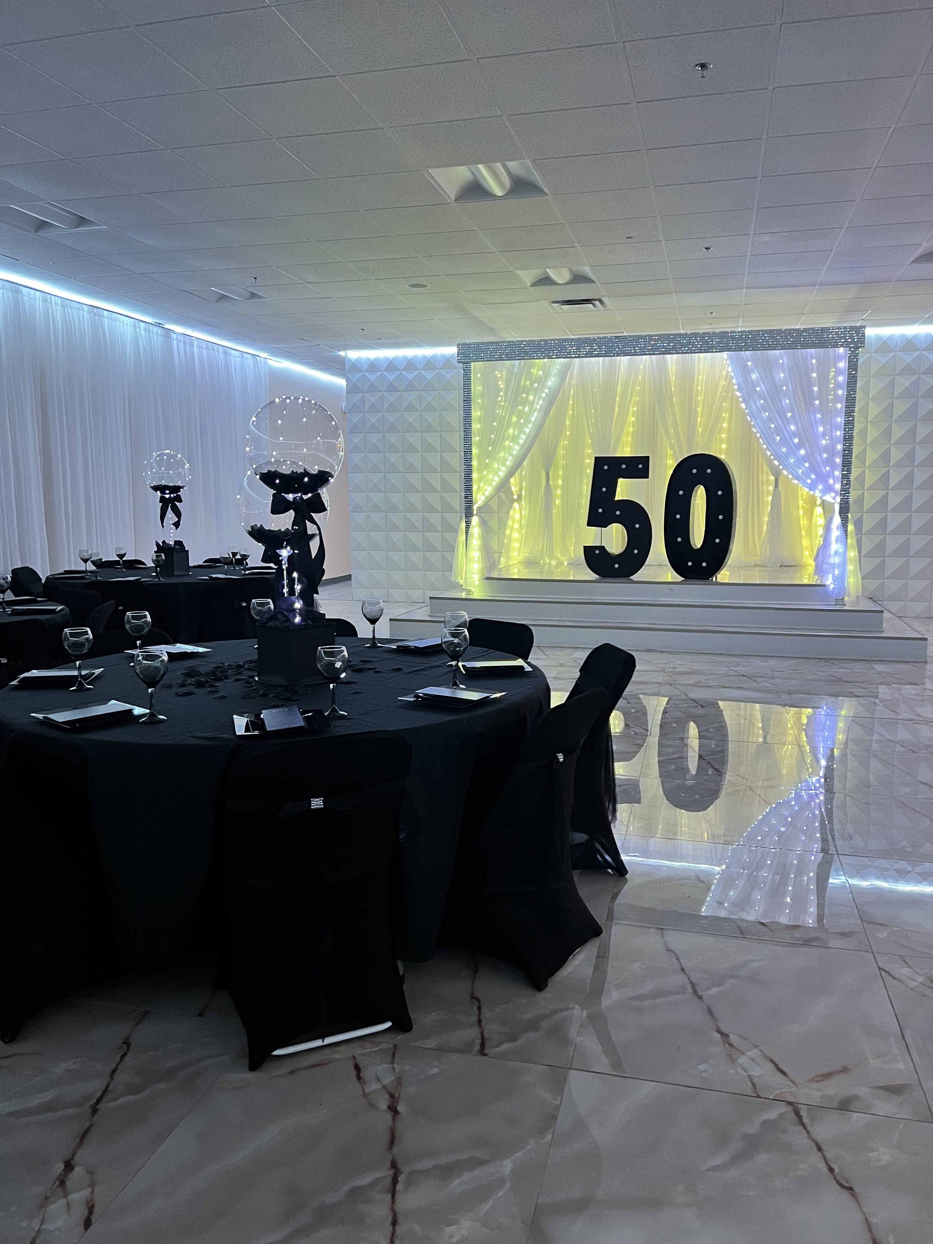 An elegant event space decorated for a 50th celebration with black tablecloths, wine glasses on tables, and a lit-up "50" sign on stage surrounded by white curtains and lights.