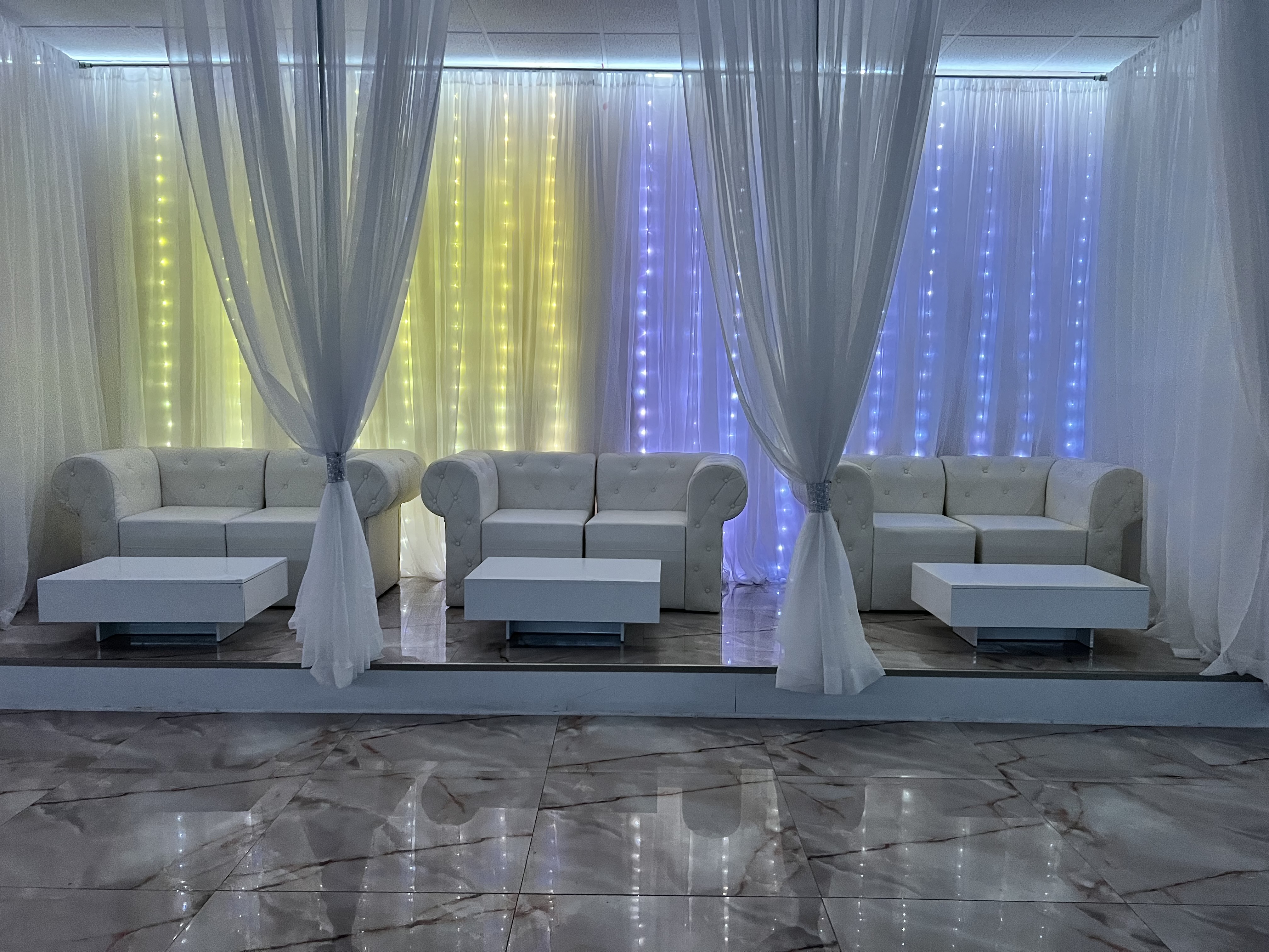 A modern event hall with white sofas and chairs set in front of a wall draped with sheer white curtains and illuminated by vertical led light strips in yellow and blue.
.