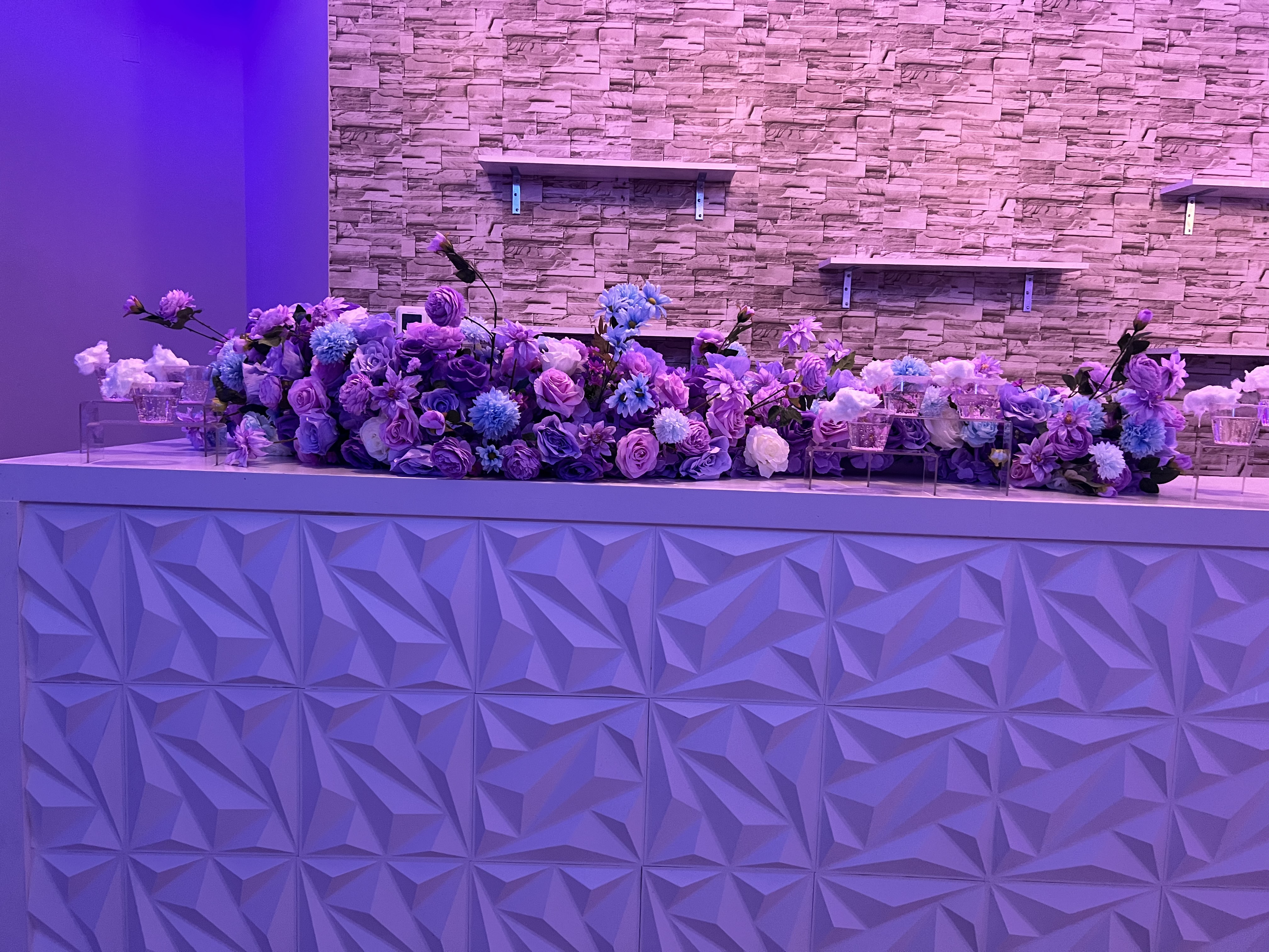 A long and elegant floral arrangement of purple and white flowers on a modern white counter, set against a textured stone wall bathed in soft purple lighting.