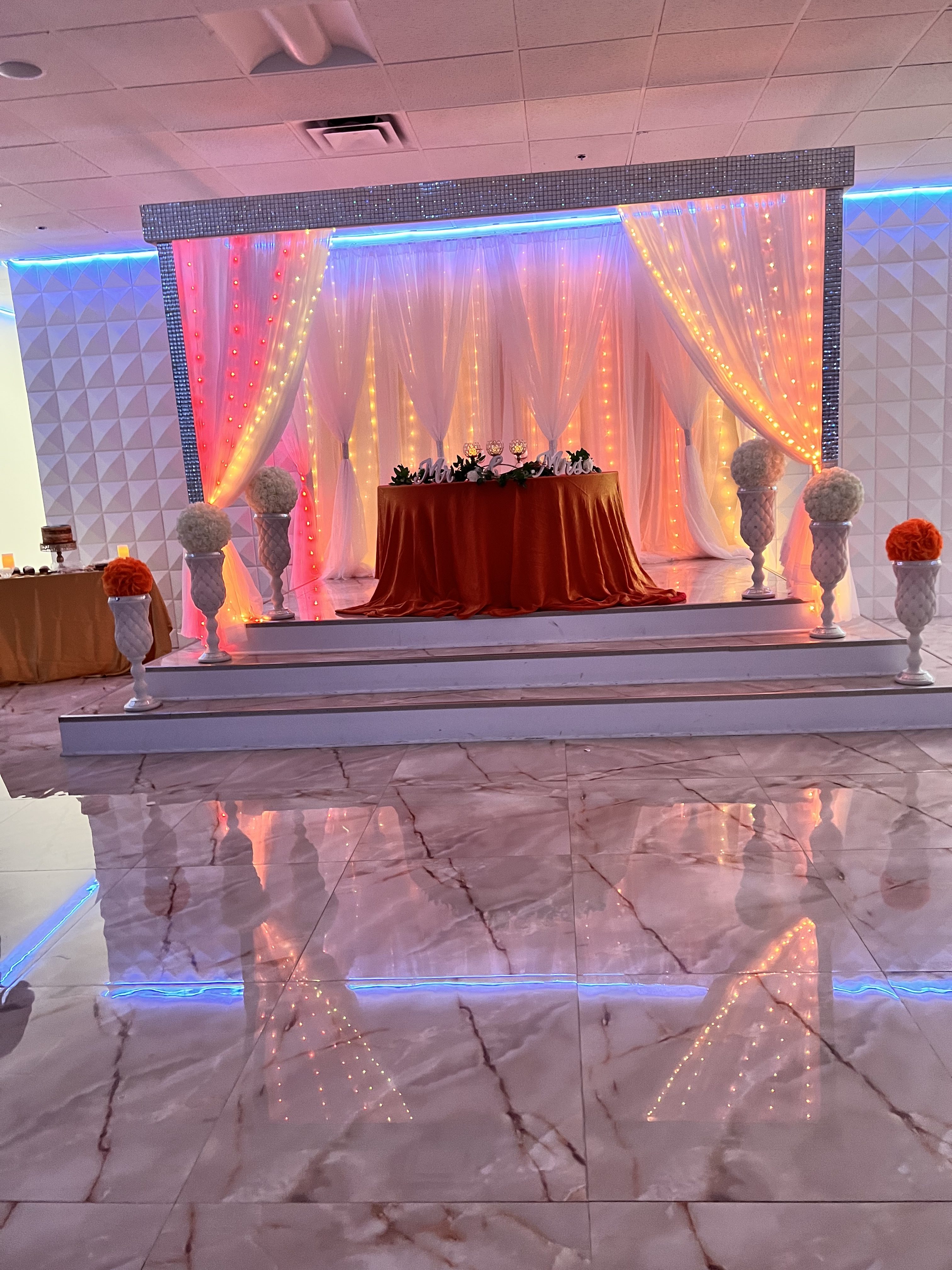 Elegant event setting with a glossy marble floor reflecting ornate decorations: a draped stage with vibrant orange and pink curtains, a large floral centerpiece on an elevated brown table, and symmetrically placed flower arrangements.