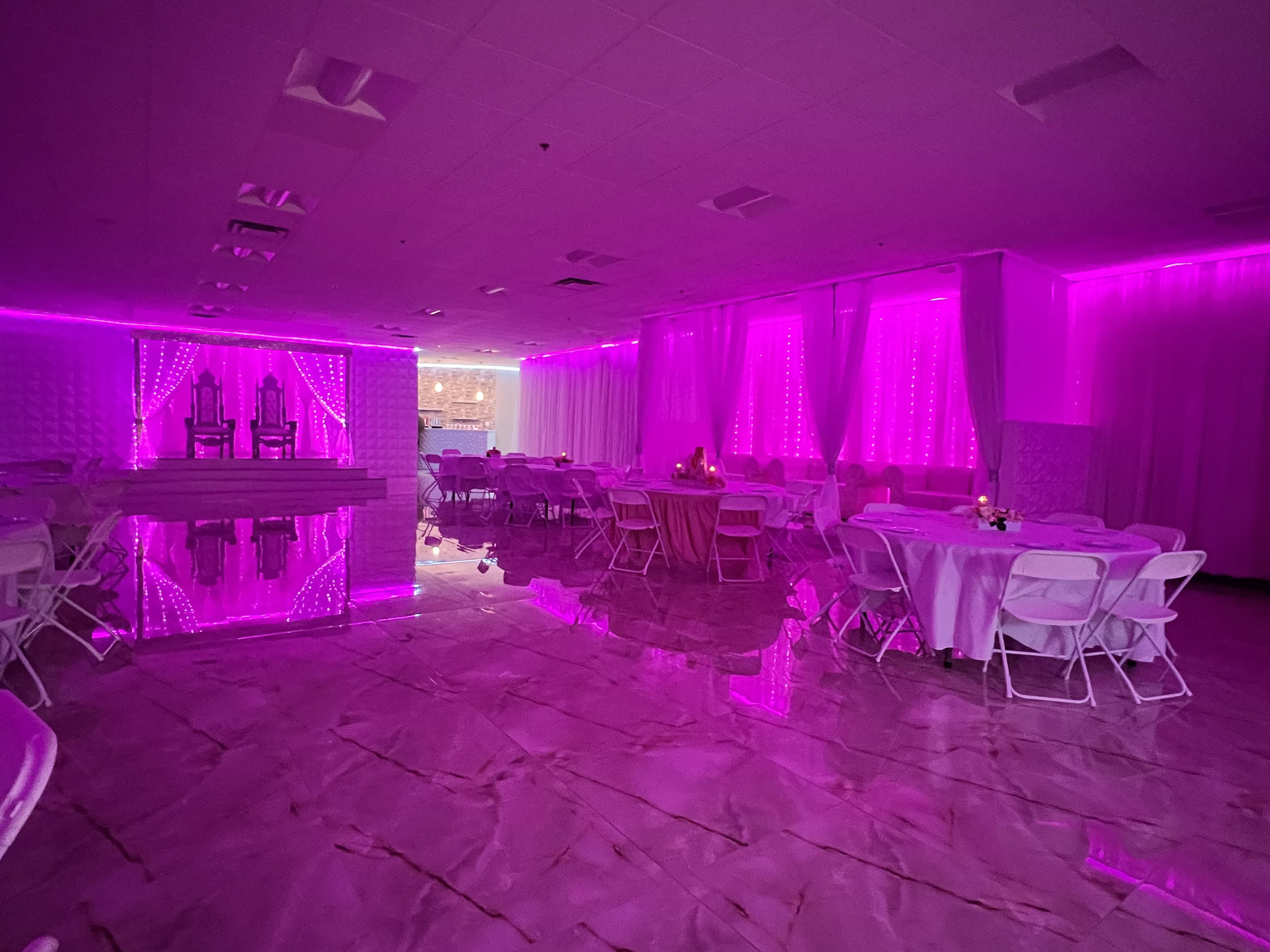 A dimly-lit event hall with violet lights, including glowing ceiling lights and neon accents along walls. reflective floor surfaces shine under white draped tables and chairs.