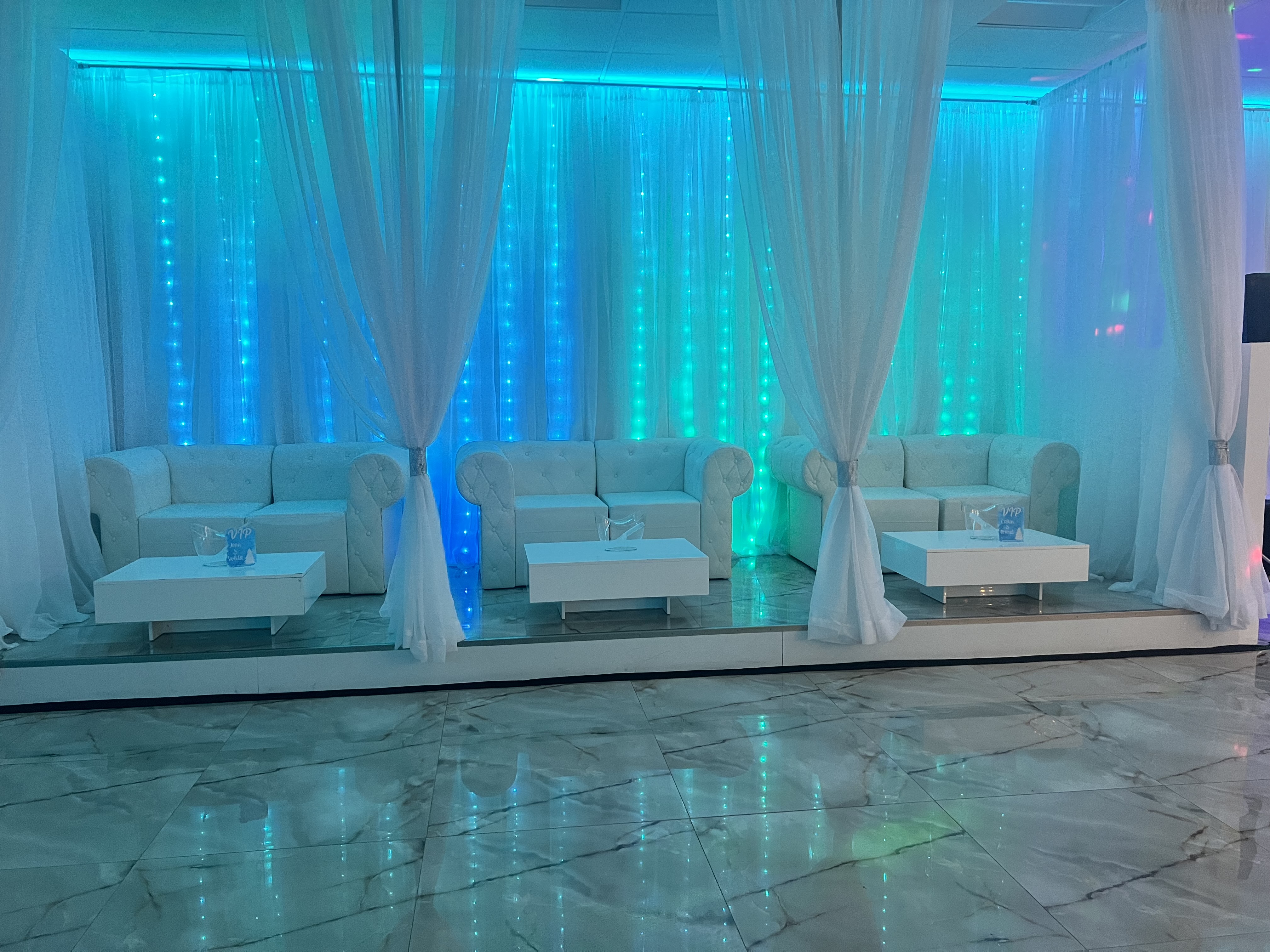 An elegant event hall featuring a long white sofa, matching tables, and draped curtains, all illuminated by a vibrant blue led light wall.