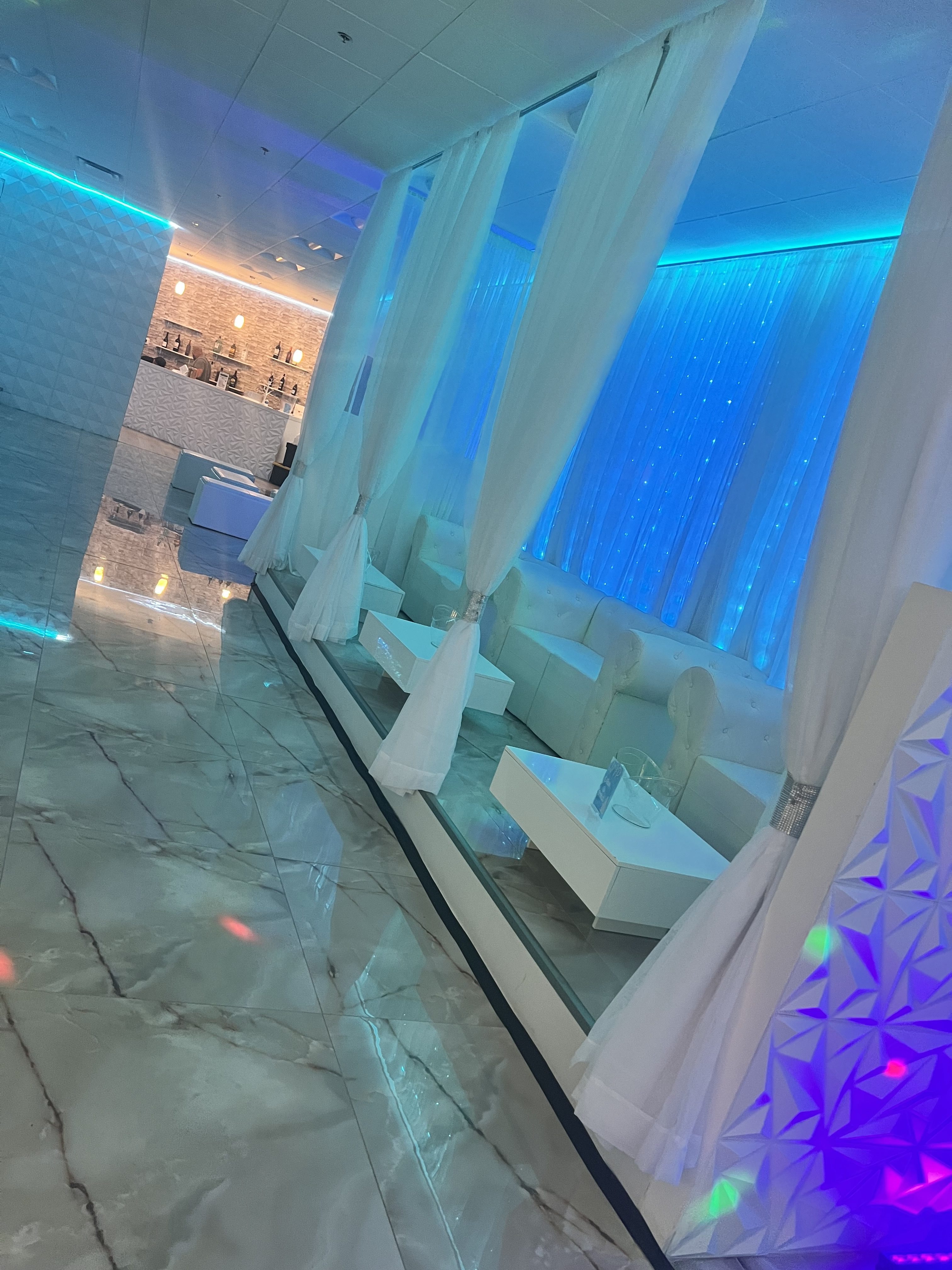 A modern lounge with marble floors, white furnishings, sheer curtains, and blue lighting creating a serene ambiance. reflective surfaces and a patterned wall accentuate the stylish interior.