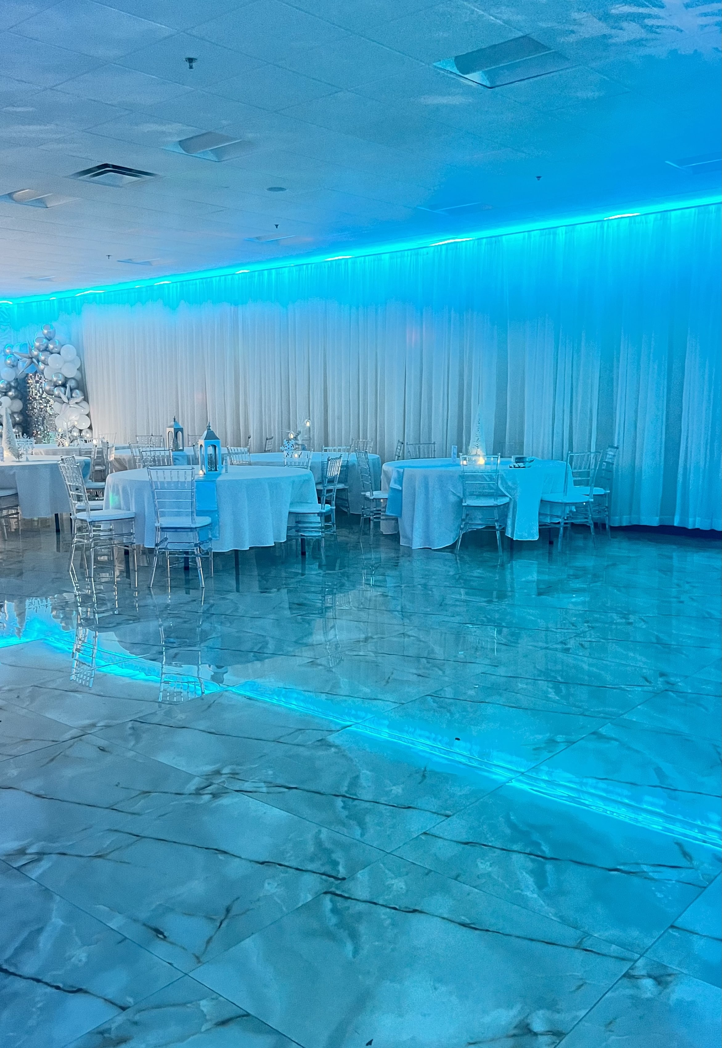 A banquet hall with marble flooring and blue lighting along the walls. tables are set with white linens and clear chairs are arranged around them.