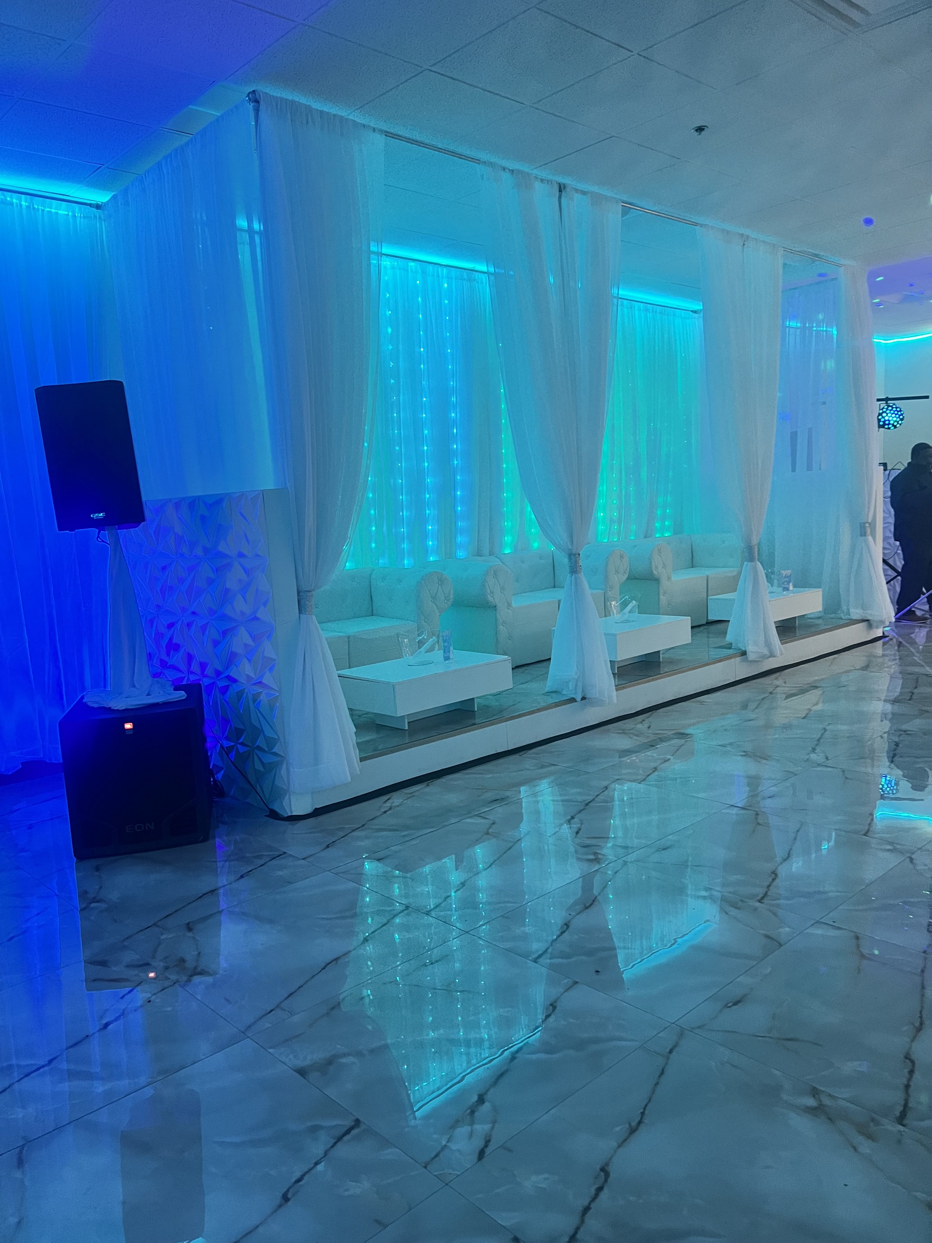 An event space illuminated in blue light with sheer white curtains, white sofas, and reflective marble floors, creating a serene, modern ambiance.