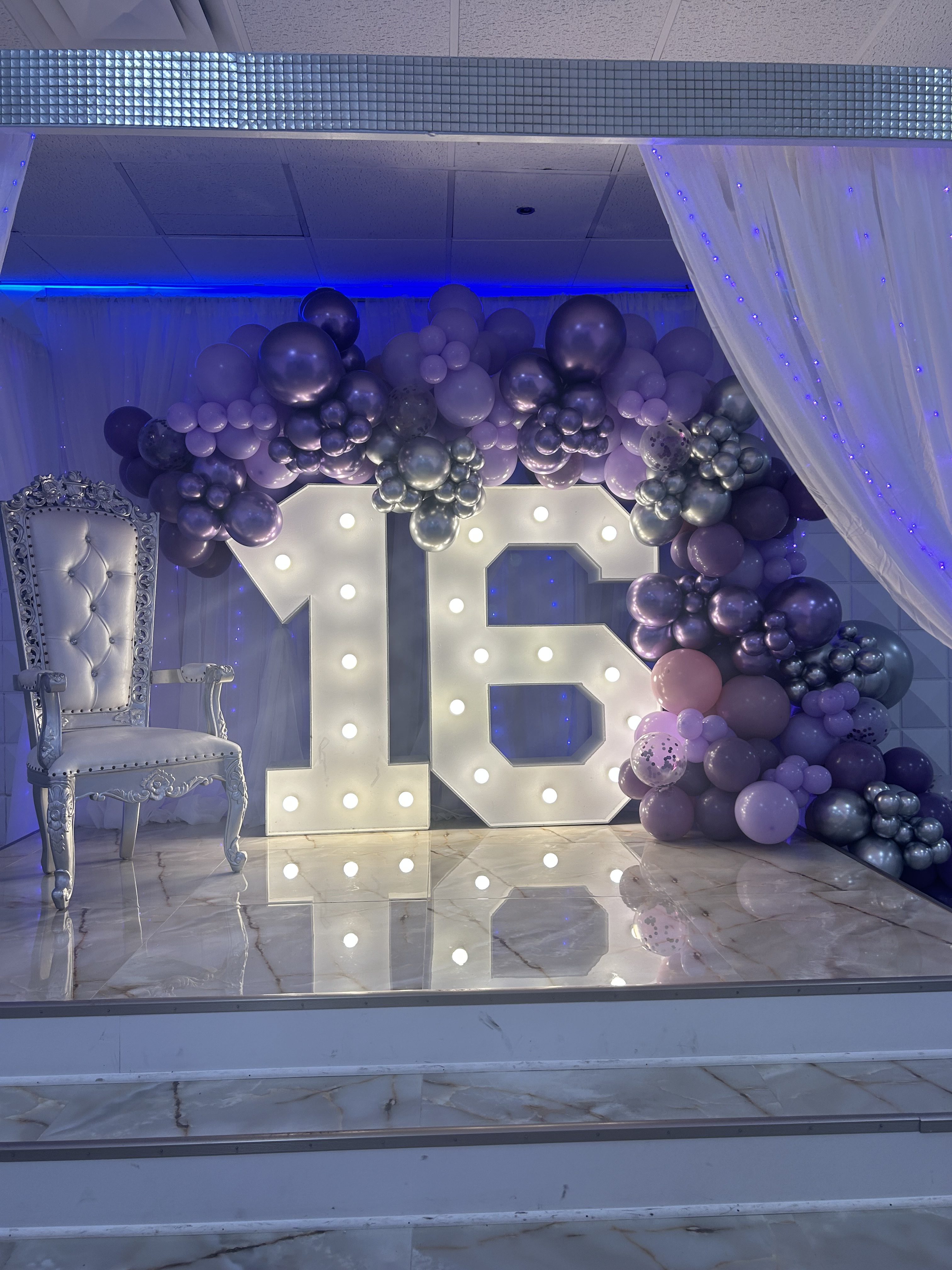 An elegant quinceañera setup featuring a large, illuminated "15" sign decorated with clusters of purple and silver balloons, next to an ornate white chair, all under soft lighting and draped fabric.