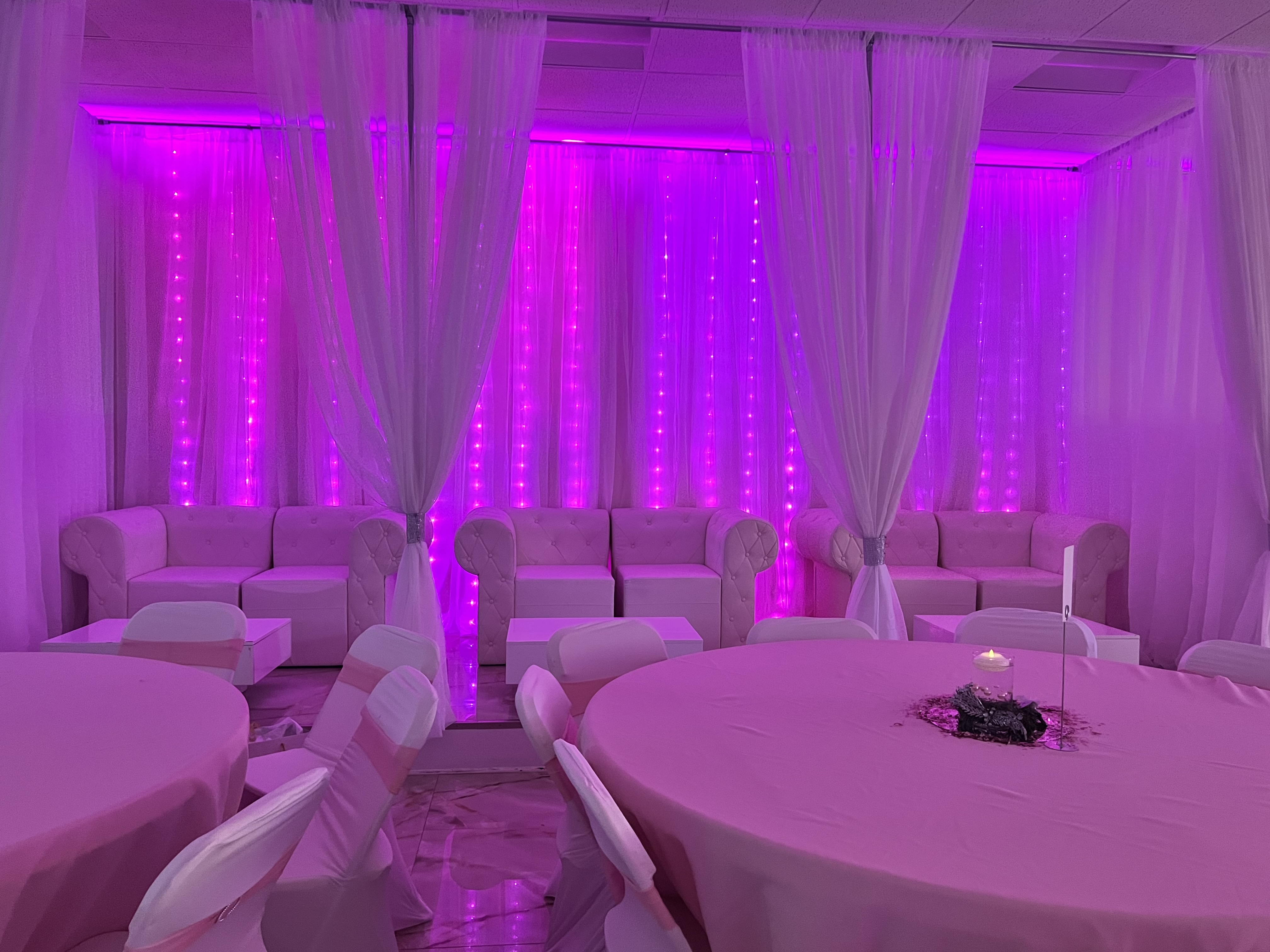 A banquet hall with pink lighting, sheer white curtains, and strands of led lights against the wall. round tables covered with pink tablecloths and white chairs arranged around.