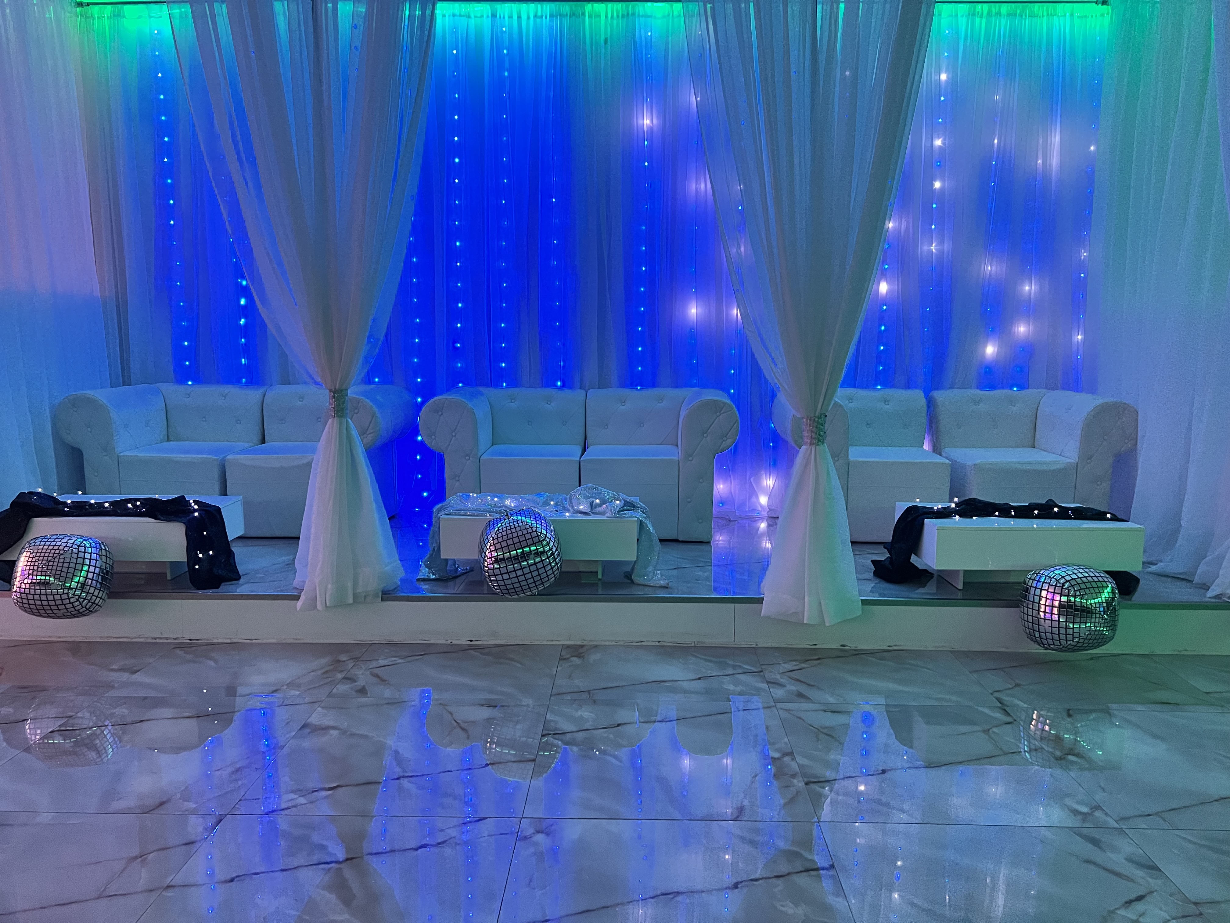 An event venue elegantly decorated with white sofas and sheer curtains. the blue-lit backdrop with twinkling lights adds a magical ambiance. crystal clear vases and mirror-like boxes reflect on the glossy floor.