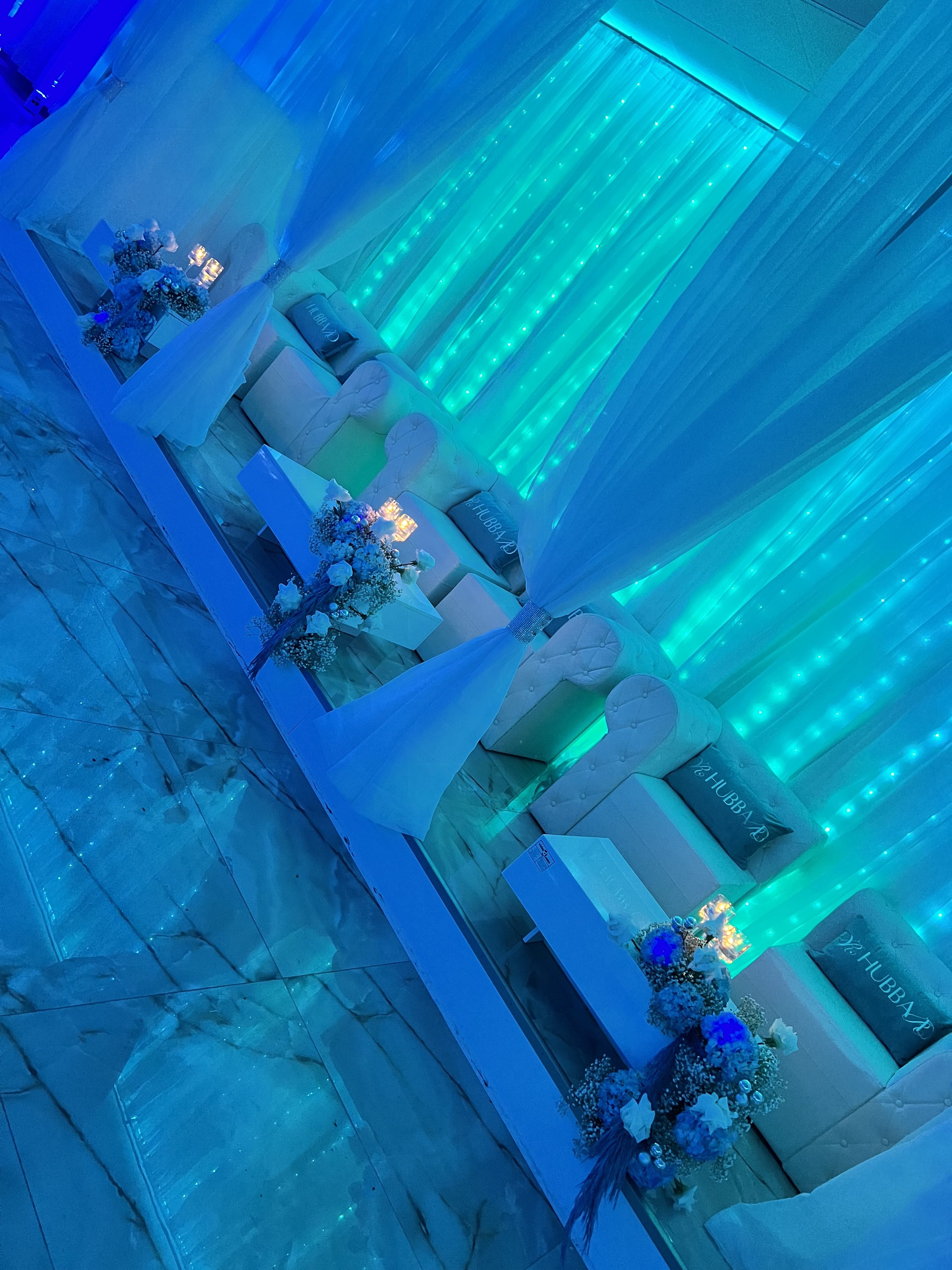 A modern event space with sleek white sofas adorned with blue and white floral arrangements, set against a wall illuminated by vertical blue neon lights.