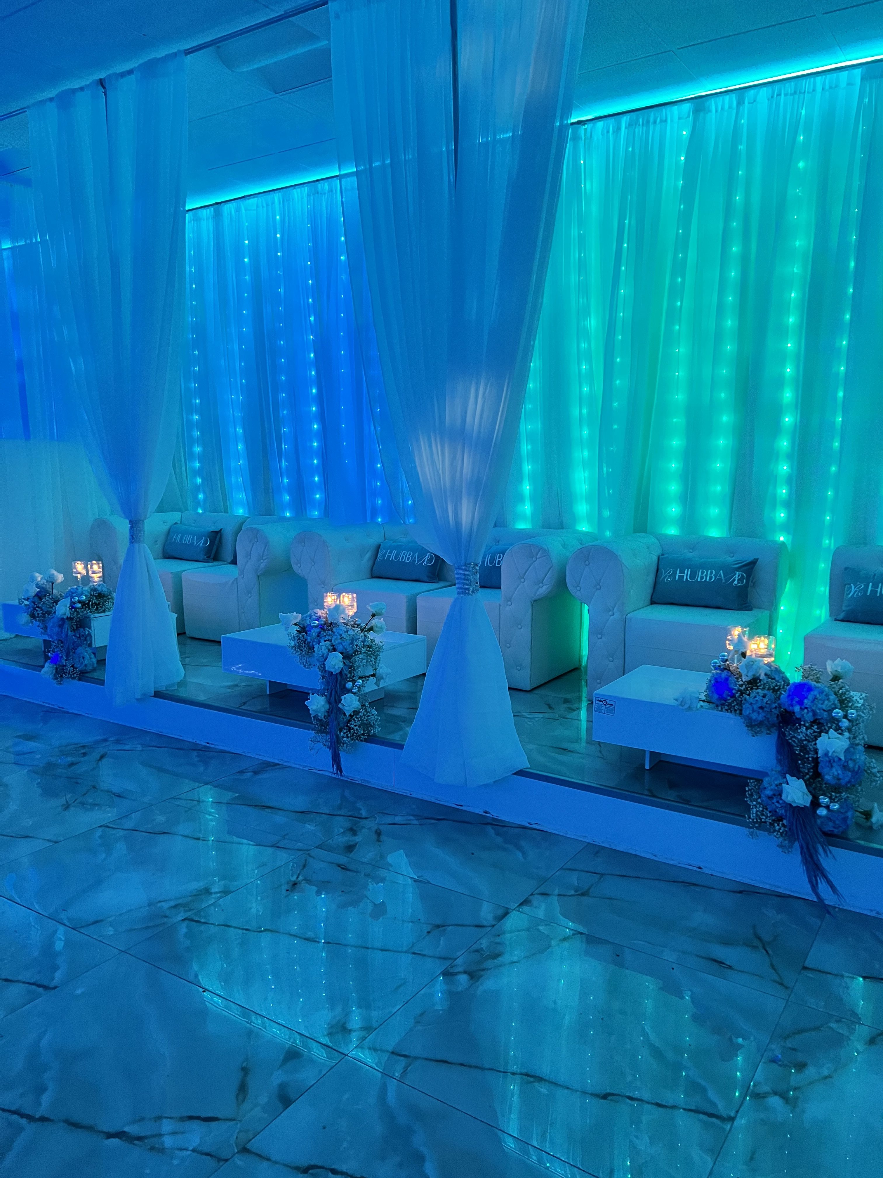 Elegant event setting with soft white sofas, floral arrangements on low tables, and a glowing blue led backdrop behind sheer white curtains, creating a serene ambiance.