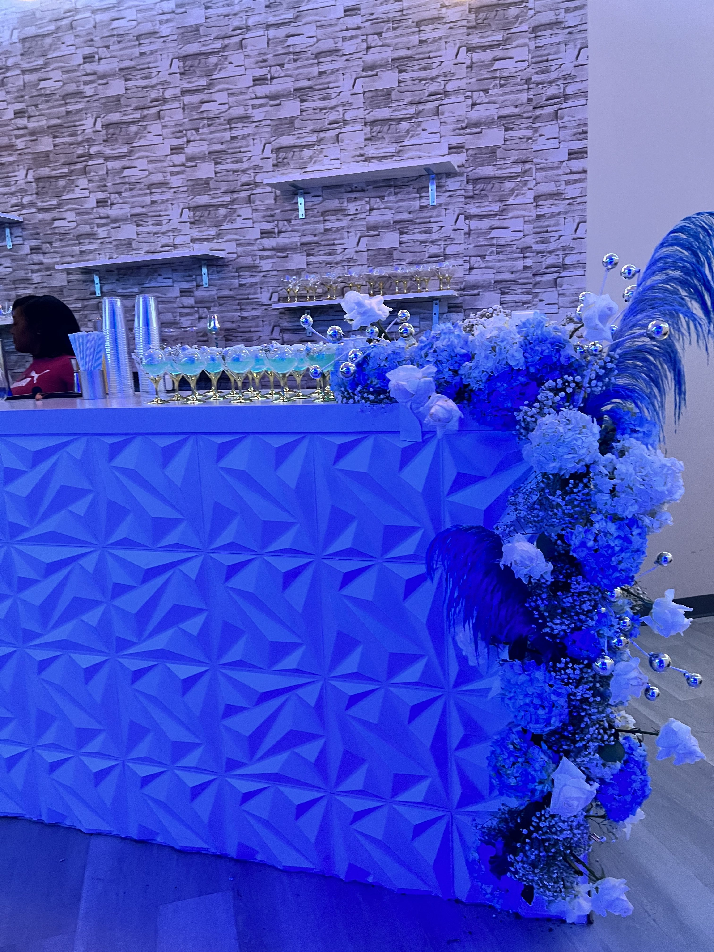 A reception desk adorned with vibrant blue and white flowers, under blue lighting, against a textured stone wall backdrop. a staff member is present behind the desk.