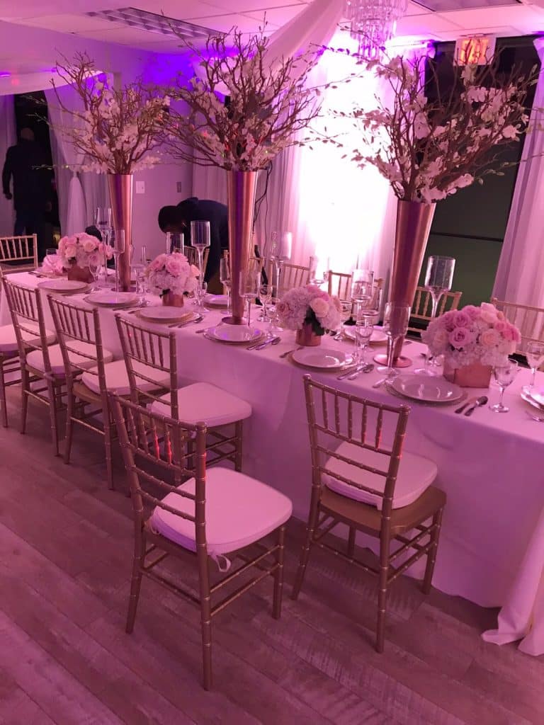 Elegant wedding reception setup featuring tables with white linens, floral centerpieces in tall vases, gold chairs, and soft pink lighting.