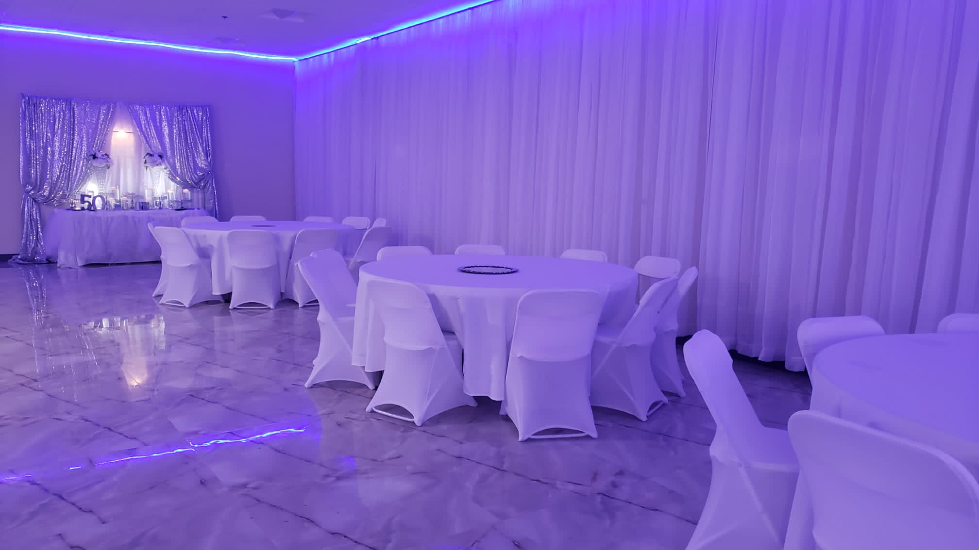 A banquet hall with purple ambient lighting, featuring round tables draped in white tablecloths and white chairs. a decorated backdrop with shimmering curtains marks the celebration area.