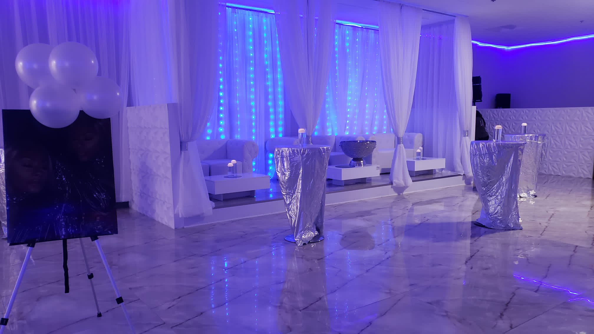 An event hall decorated in blue and white, featuring glowing blue lights, white draped curtains, silver high-top tables, and white lounge furniture. balloons and large photo panels add to the decor.