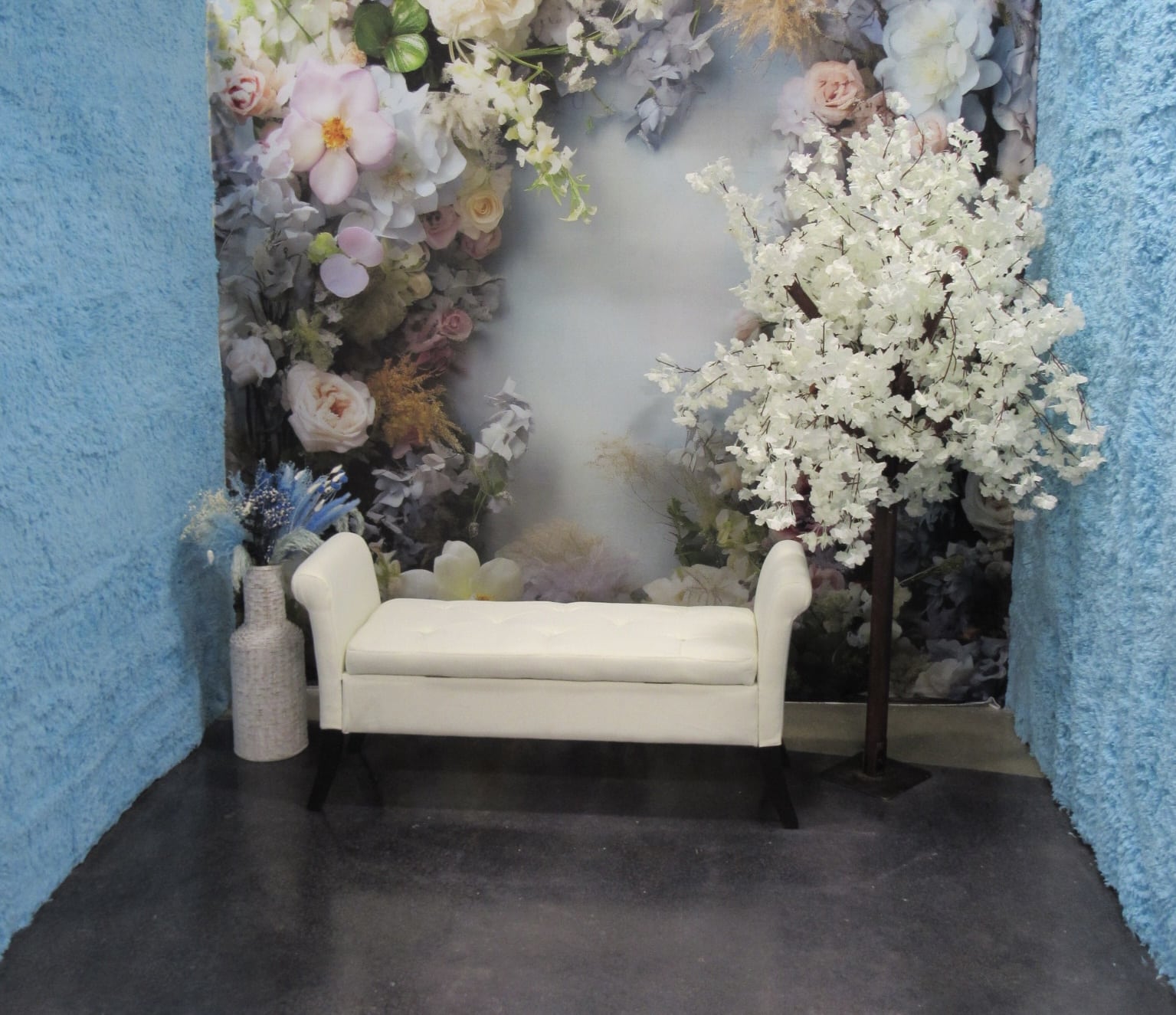 A cozy corner with a small white sofa against a backdrop of lush, assorted artificial flowers in soft colors and a white floral tree beside it, surrounded by textured blue walls.
