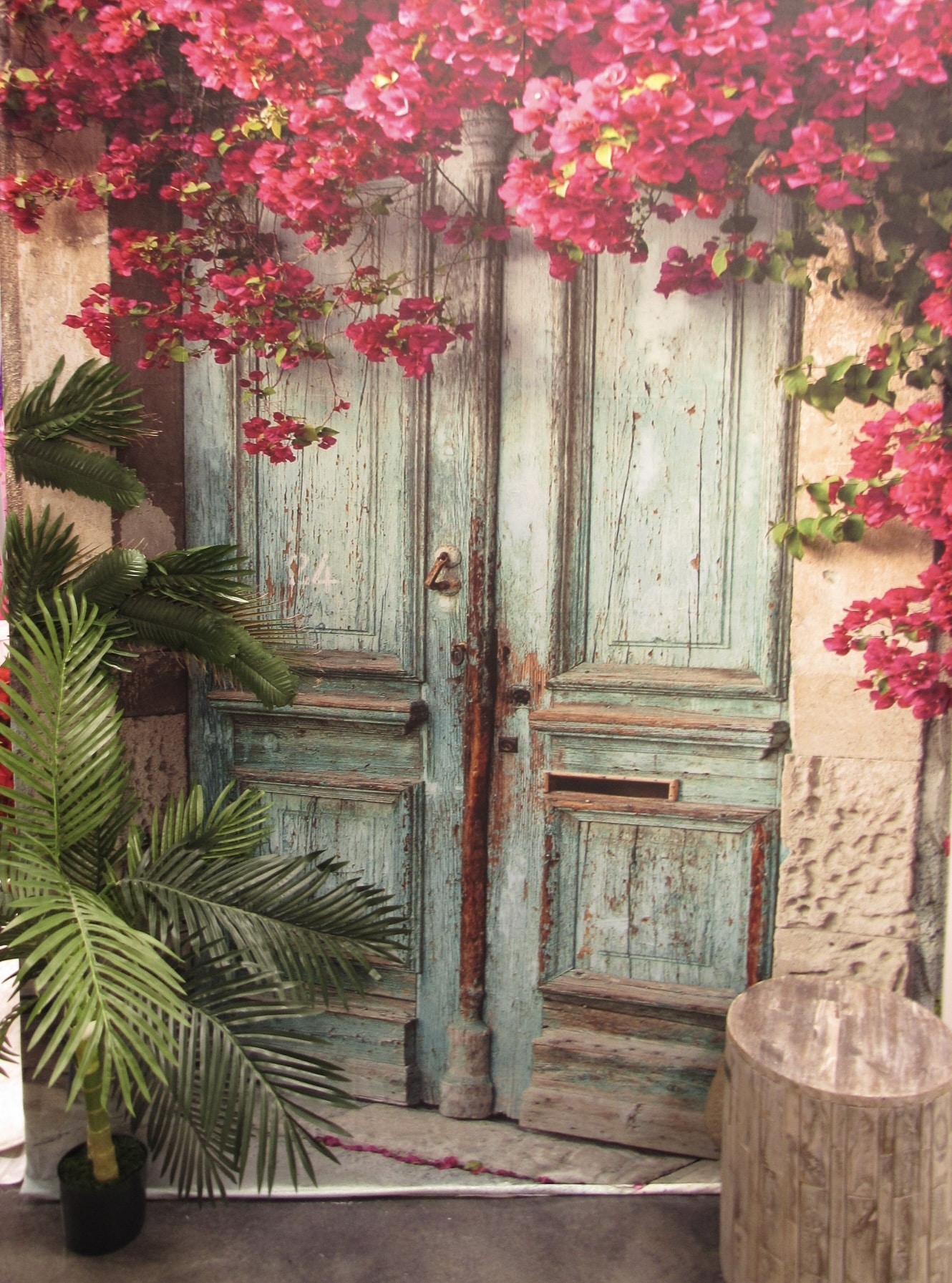 A rustic weathered turquoise door with ornate metal handles, framed by lush pink bougainvillea blossoms and a green plant to the left, set in an old stone wall.