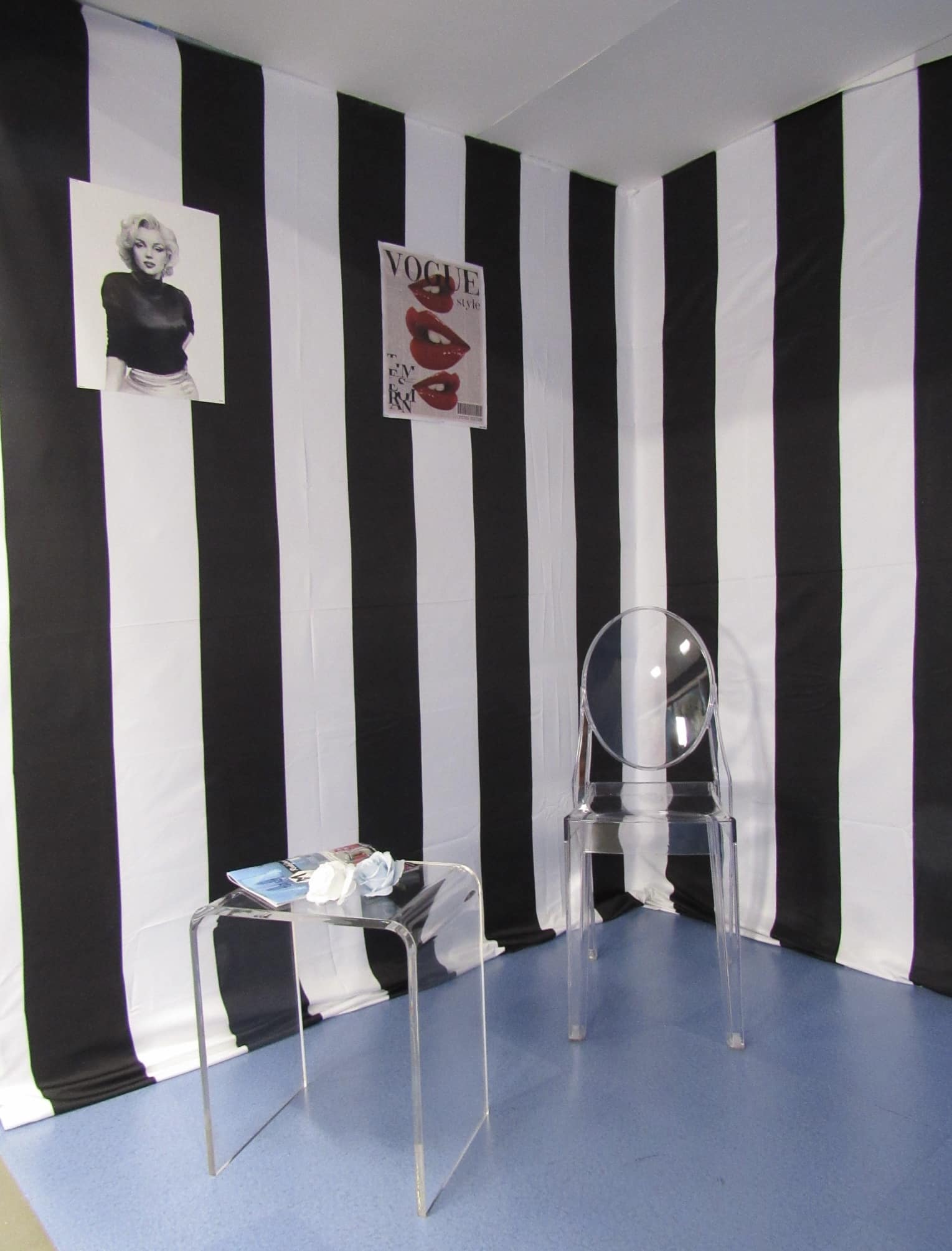 A stylish corner with black and white striped walls, featuring posters of marilyn monroe and a vogue cover. there's a transparent bubble chair, a small glass table with magazines, under soft lighting.
