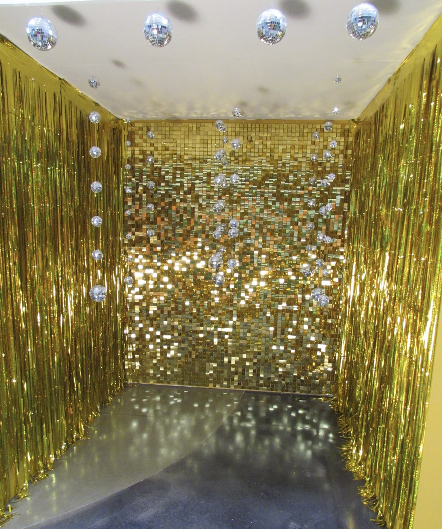 A vibrant photo showcasing a corner filled with golden, shimmering foil curtains and multiple disco balls hanging from the ceiling, reflecting light on a gray floor.
